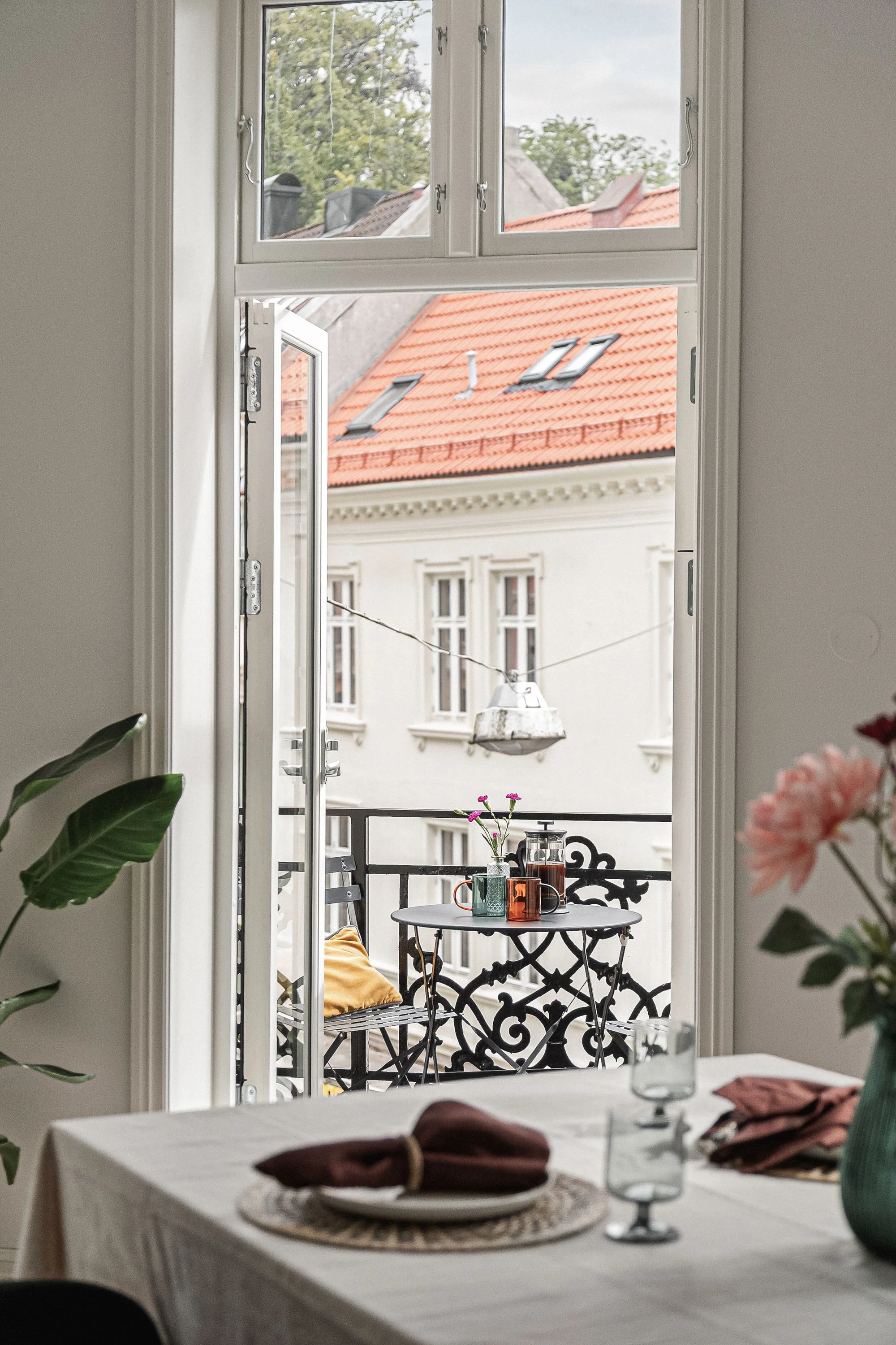 Et spisebord dekket med tallerkener, glass og duk, sett innenfor et rom med åpent vindu mot en balkong. På balkongen er det et lite rundt bord med blomster og kopper, omgitt av et metallrekkverk, med en bynært bygårdsfasade og tak med taklys i bakgru