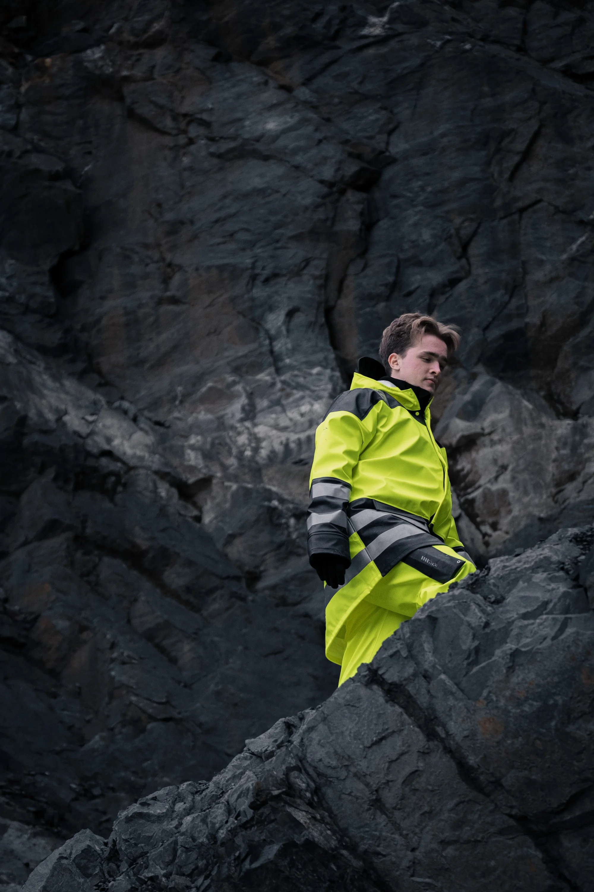 En mann i en neon gul regnjakke som står på store svarte steiner i en fjell- eller klippeomgivelser.