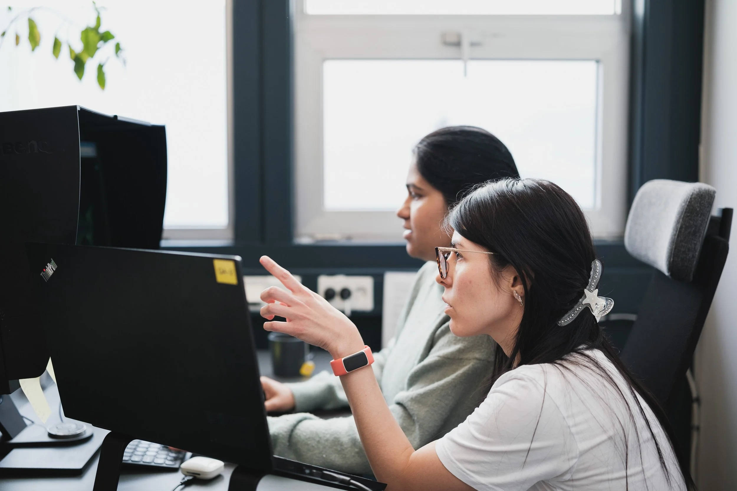 To kvinner sitter ved kontorarbeidsstasjoner og jobber på datamaskiner.