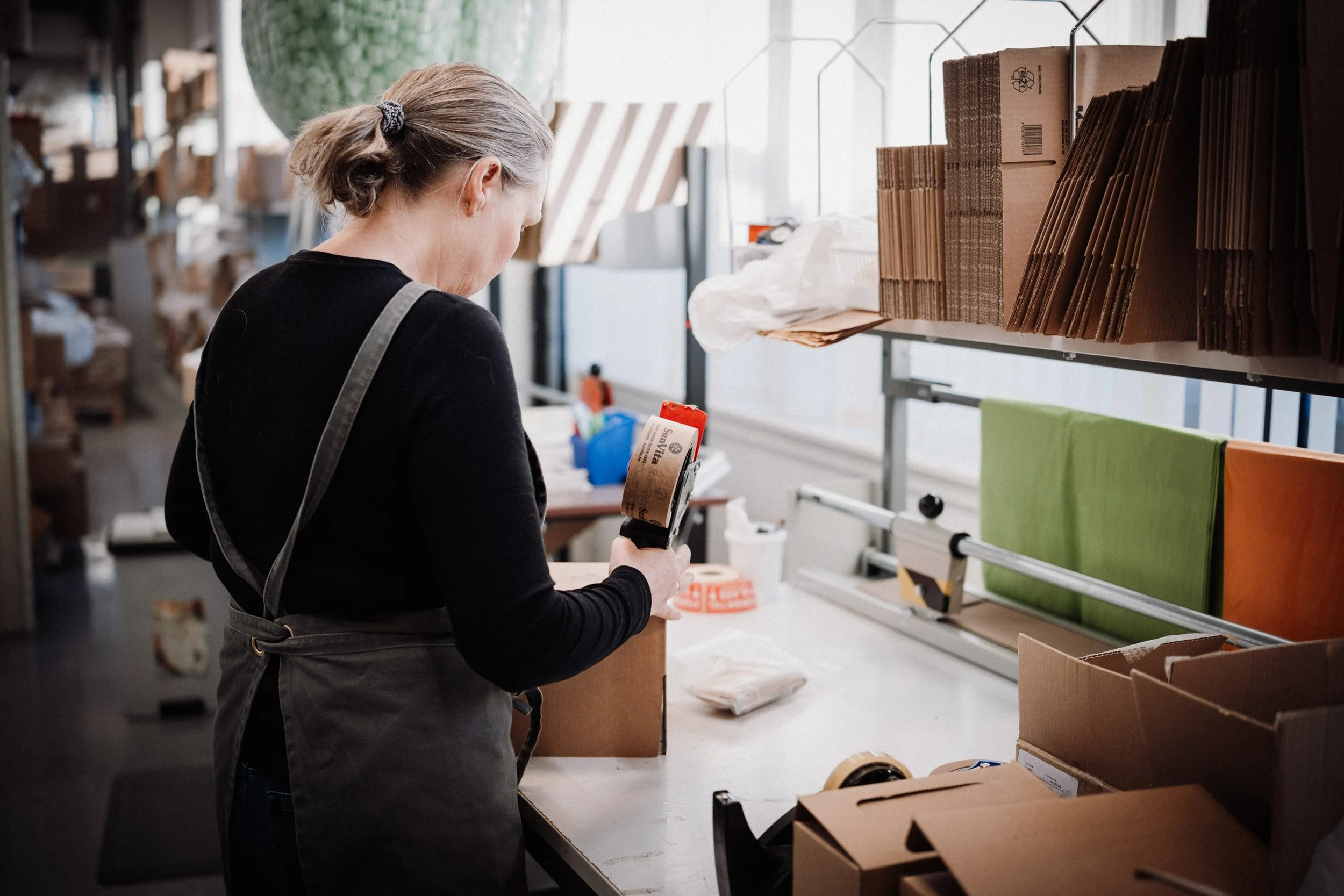 En kvinne jobber på en emballasjebedrift, pakker varer i en kasse i et industrielt miljø.