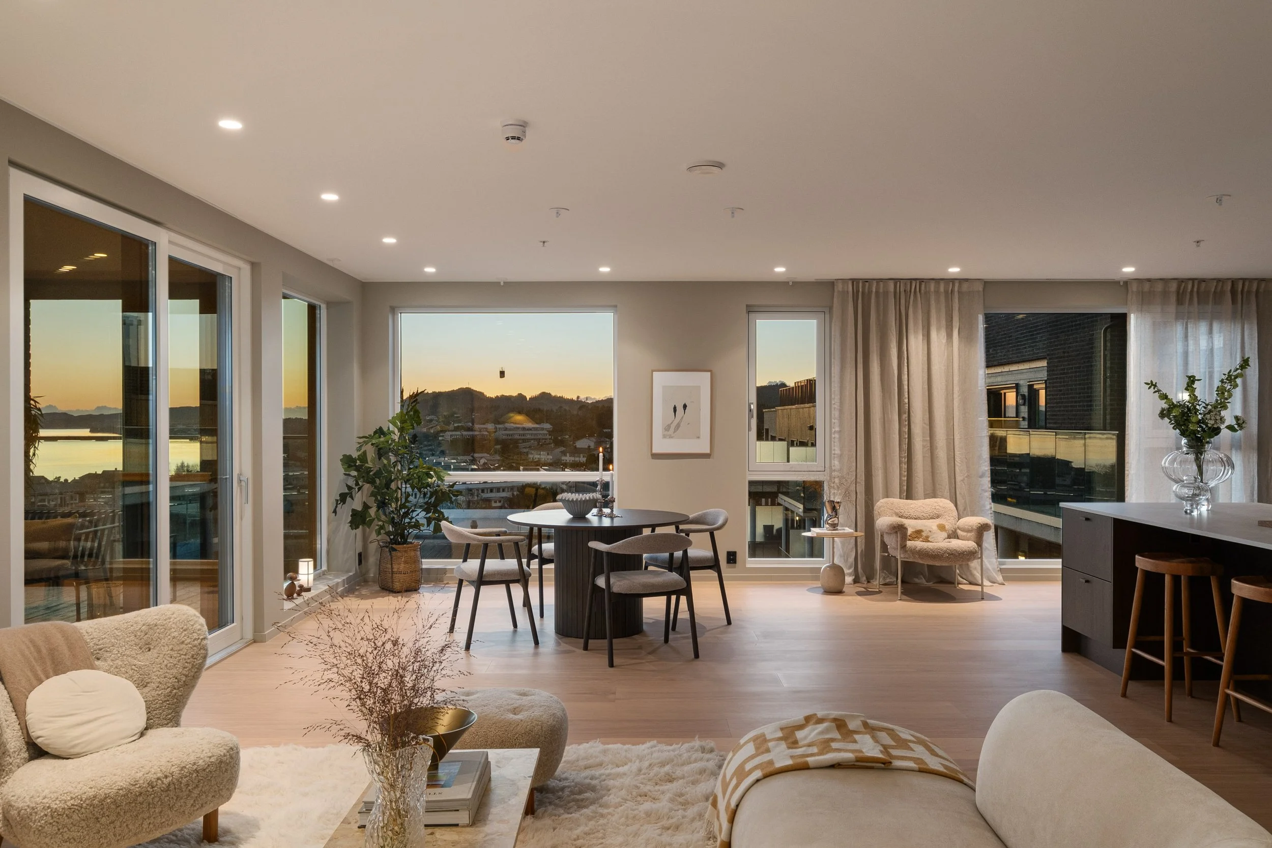 Et lyst og moderne stueområde med store vinduer som viser en bygd med vann og fjell i bakgrunnen, møbler i lyse farger, grønne planter og dekorative elementer.