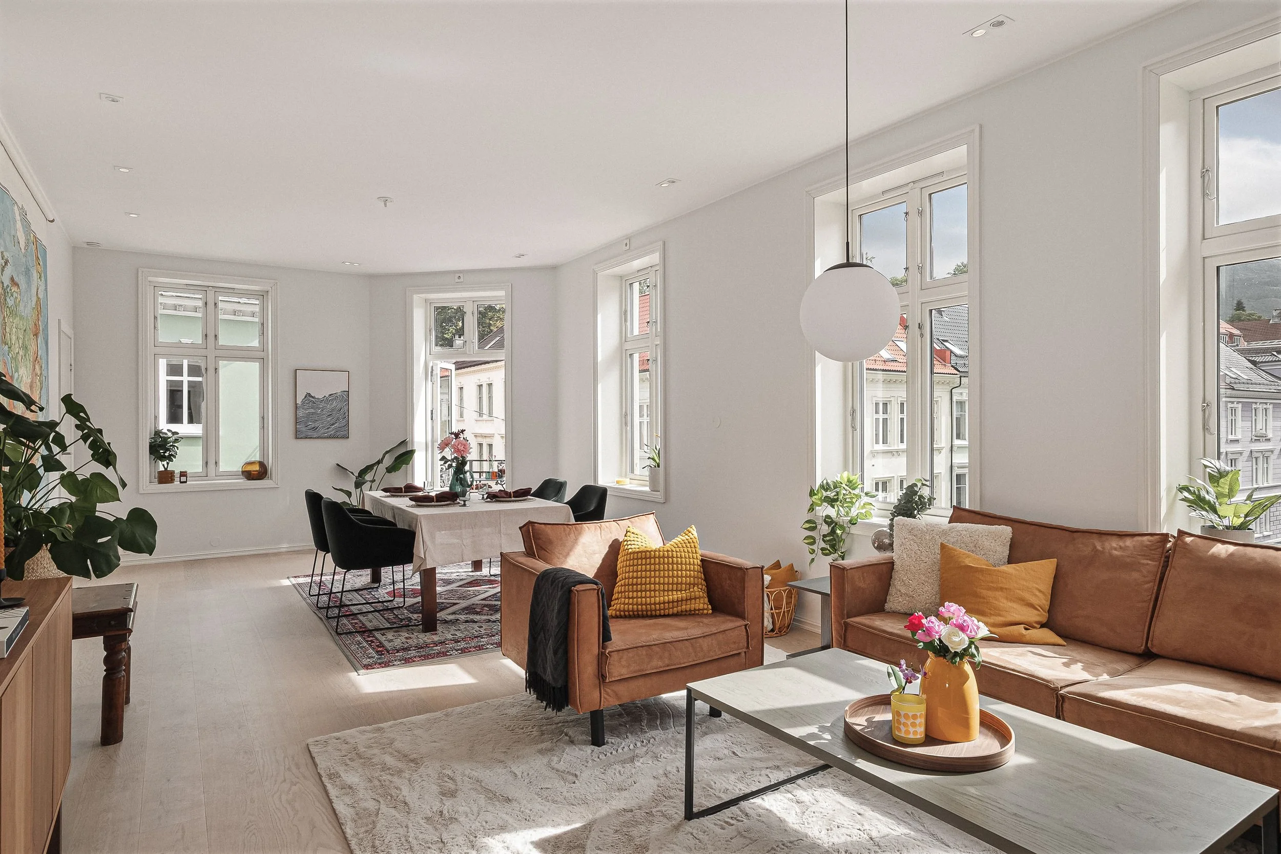 Lyse stue med store vinduer, sofa, stuebord og spisestue med spisestue, grønne planter, pyntegjenstander og veggkunst.