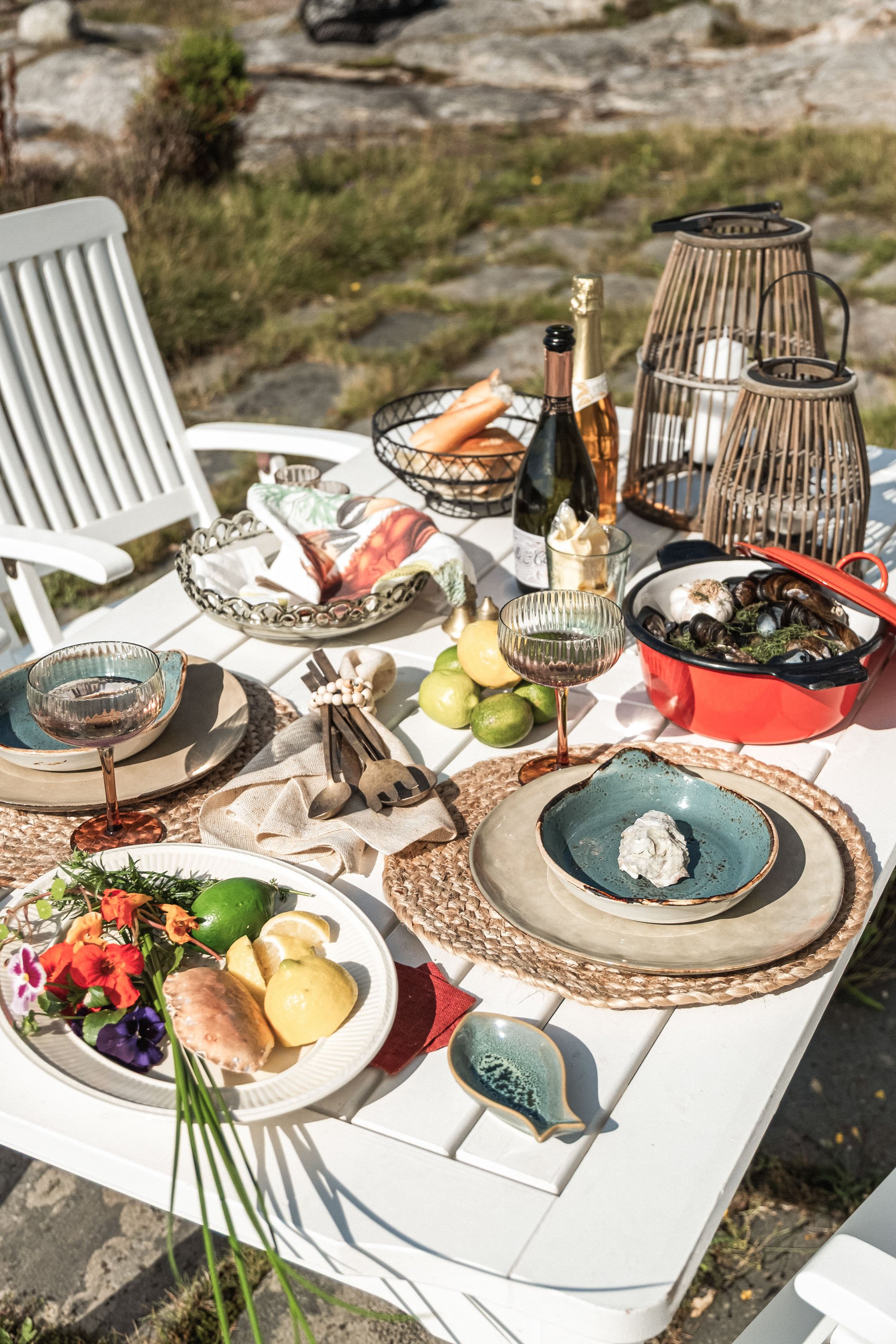 Utendørs middagsbord med tallerkener, glass, vin og mat, omgitt av kaste stoler og dekorative lanterner.