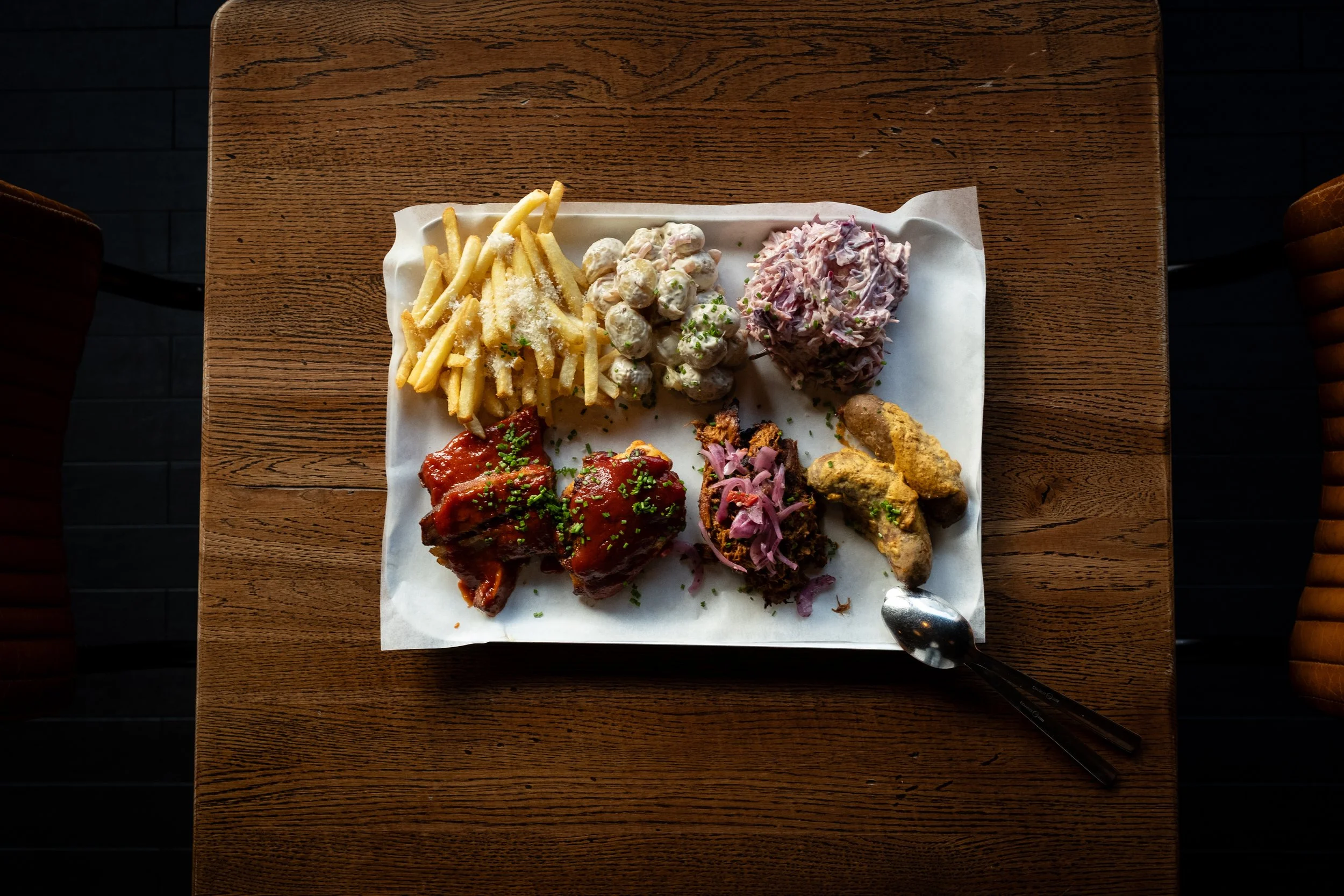 Tallrik med frityrstekt mat, pommes frites, potetsalat, kål, köttbullar med gräddsås, köttklubbor, samt två knivar och en sked på ett träbord.