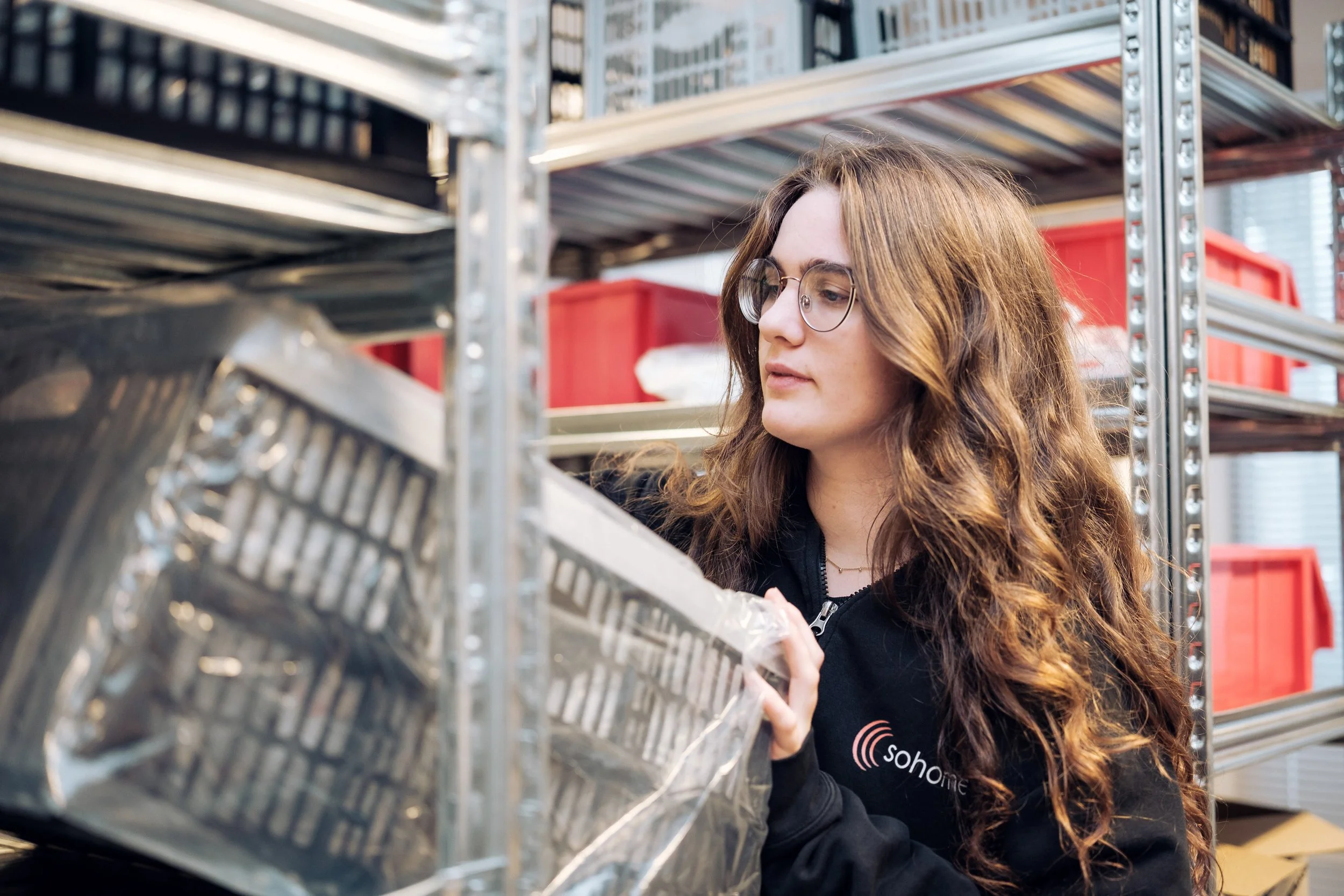 En kvinne med briller og langt, krøllete hår er i ferd med å plukke opp en metallboks fra en hylle i et lager. Hun bruker en svart jakke med logo som sier 'sohome'.