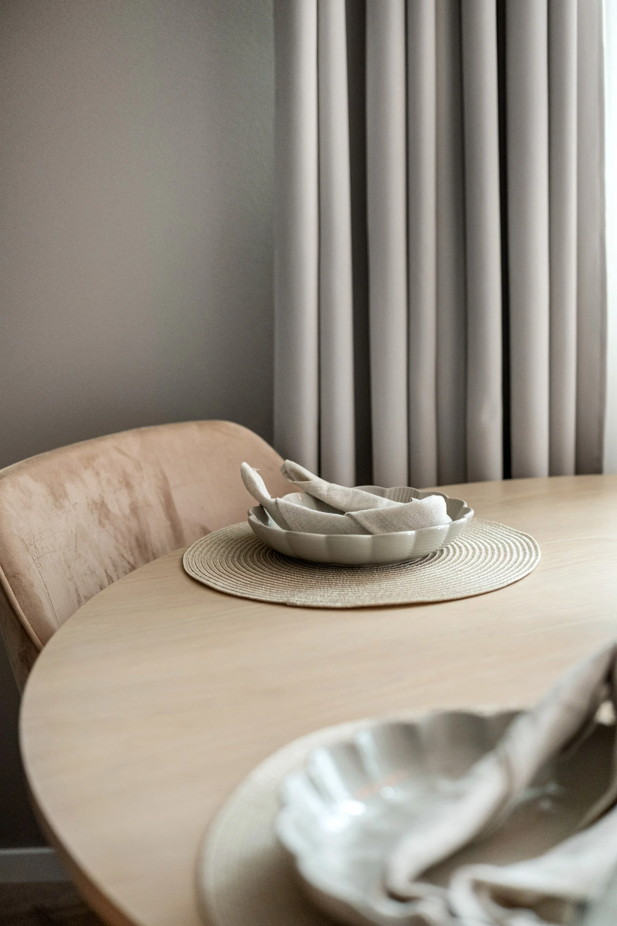 Et bord med putetrekk og servietter foran en vinduskarm med hvite gardiner, i et rom med grå vegg.