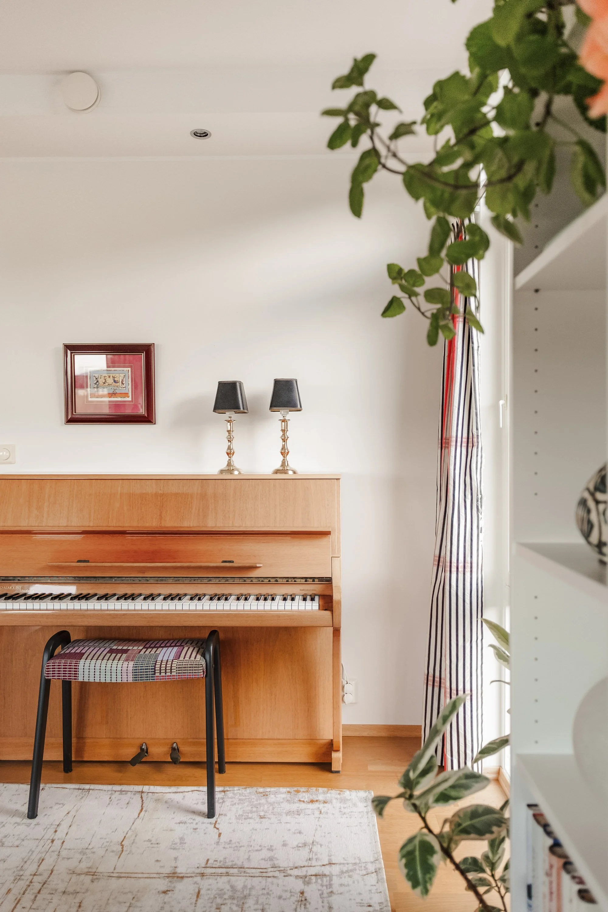 Et rom med en åpen, trehvitt elektrisk piano mot en hvit vegg med to små svarte lamper på toppen. En rødrammet kunsttrykk henger på veggen. Til høyre er det et vindu med striper på gardinen, og noen grønne planter er delvis synlige på høyre og neders