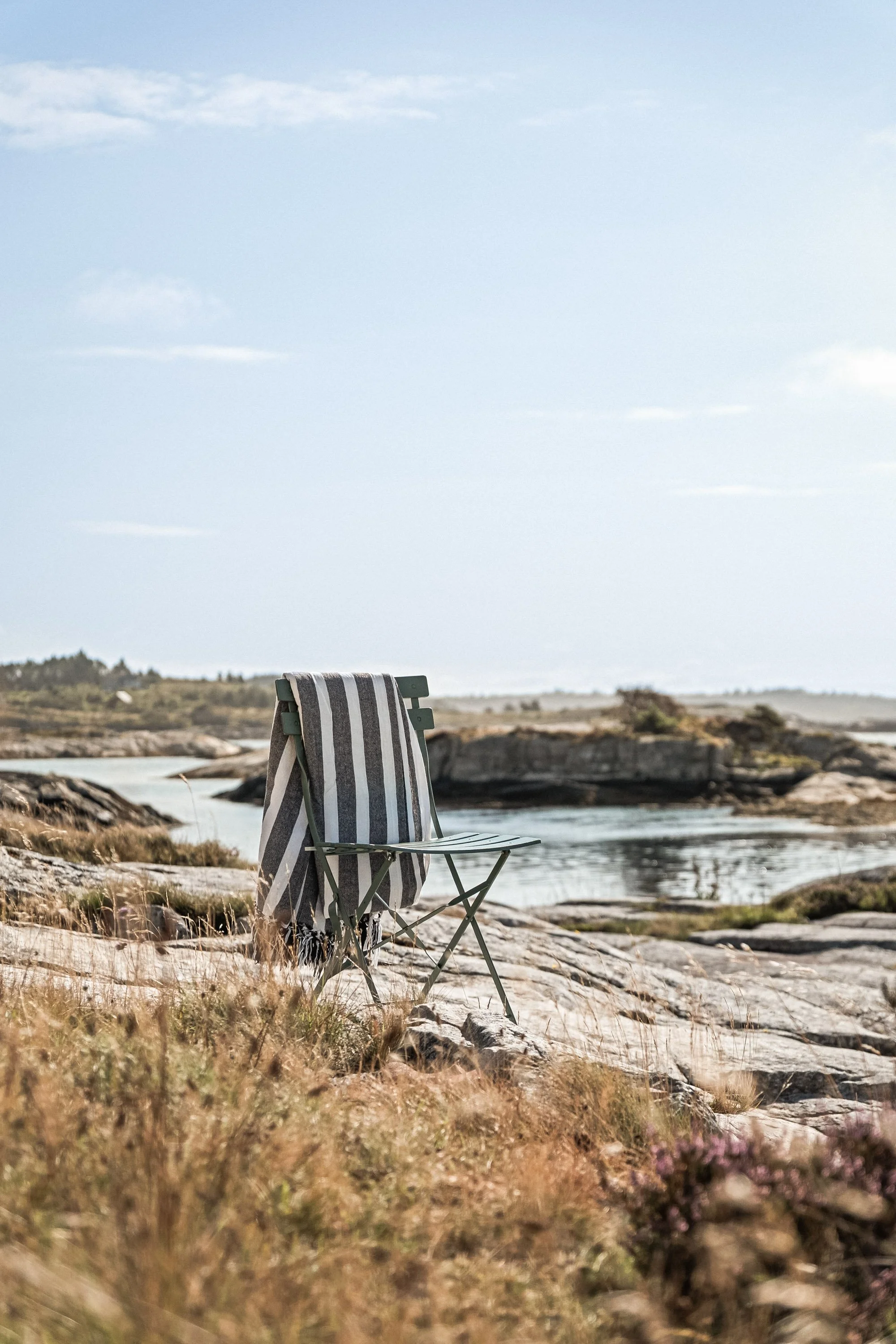 En tom campingstol med et stripe-tekstiltolld drapert over, plassert på en steinete strand med vann og lav vegetasjon i bakgrunnen, under en overfylt himmel.