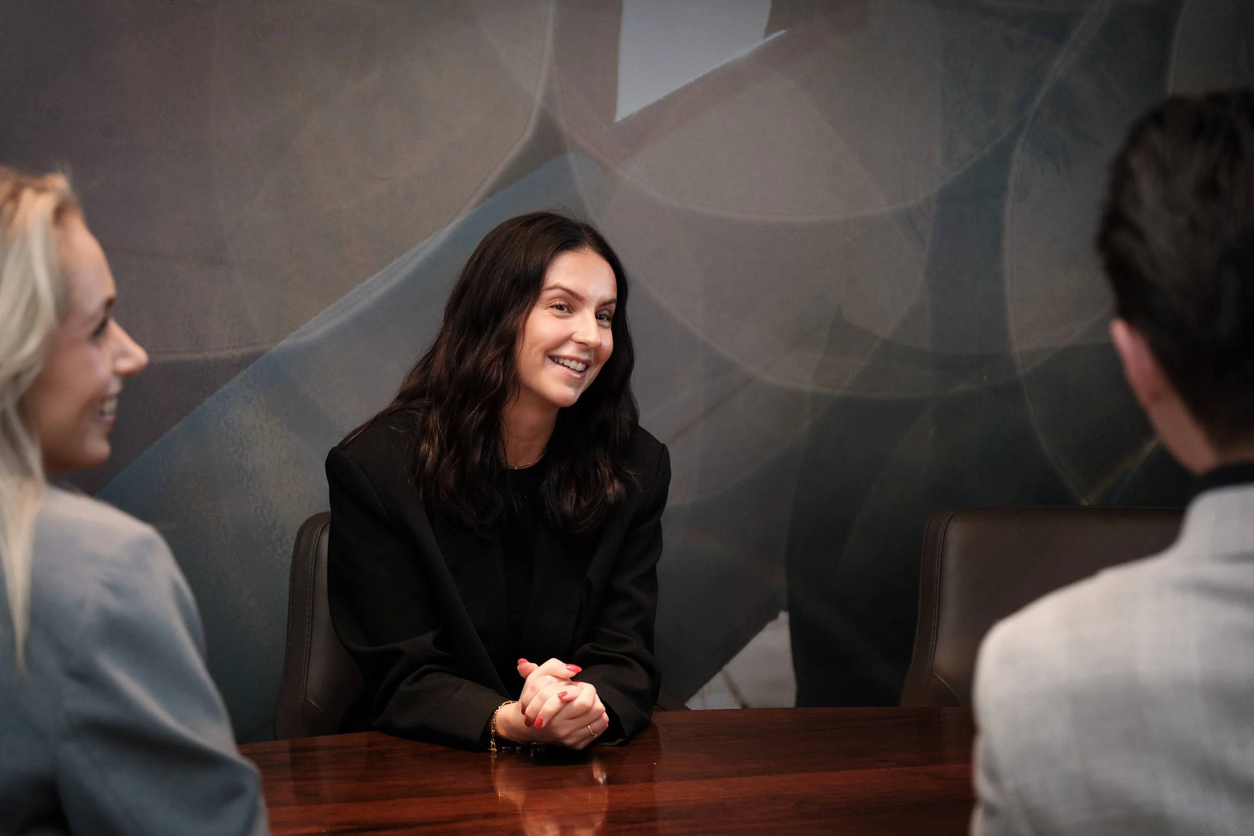 En gruppe mennesker som sitter rundt et bord i et møte. Den sentrale personen er en kvinne med mørkt, bølget hår som smiler.