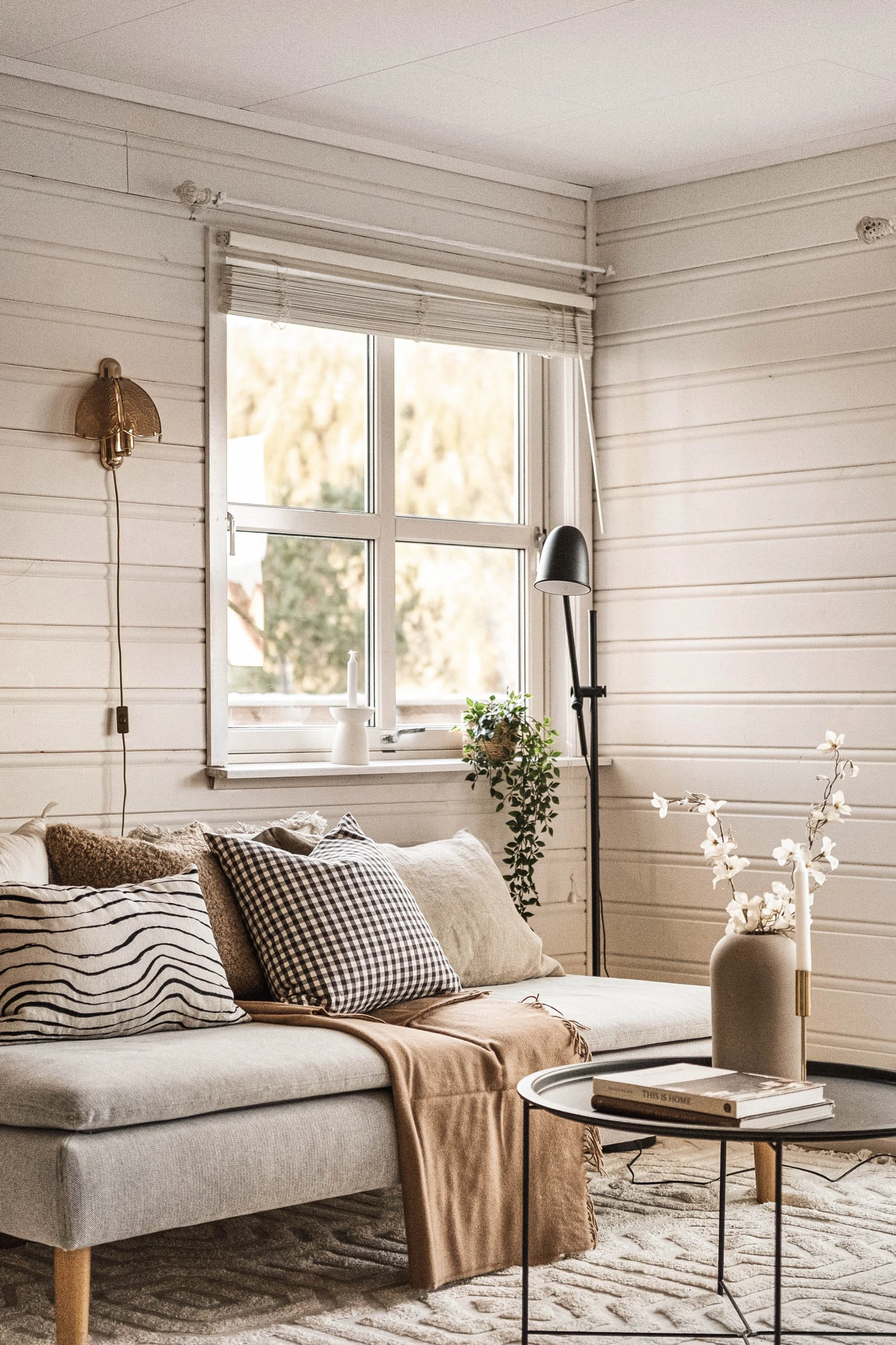 Stue med hvit vegg, sofa med puter, vindu med gardin, gulvlampe, vase med blomster, bord med bøker.