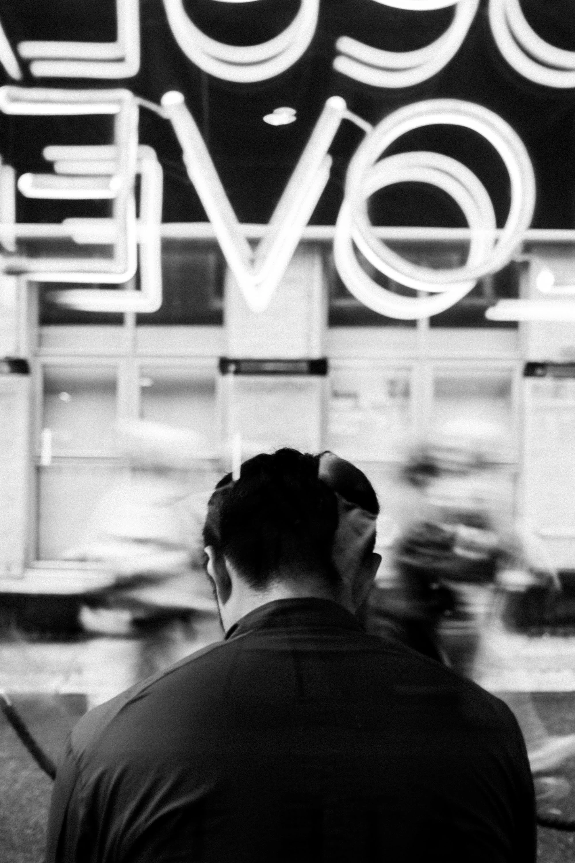 En person sett bakfra som sitter foran en butikkvindu med neon-skilt.