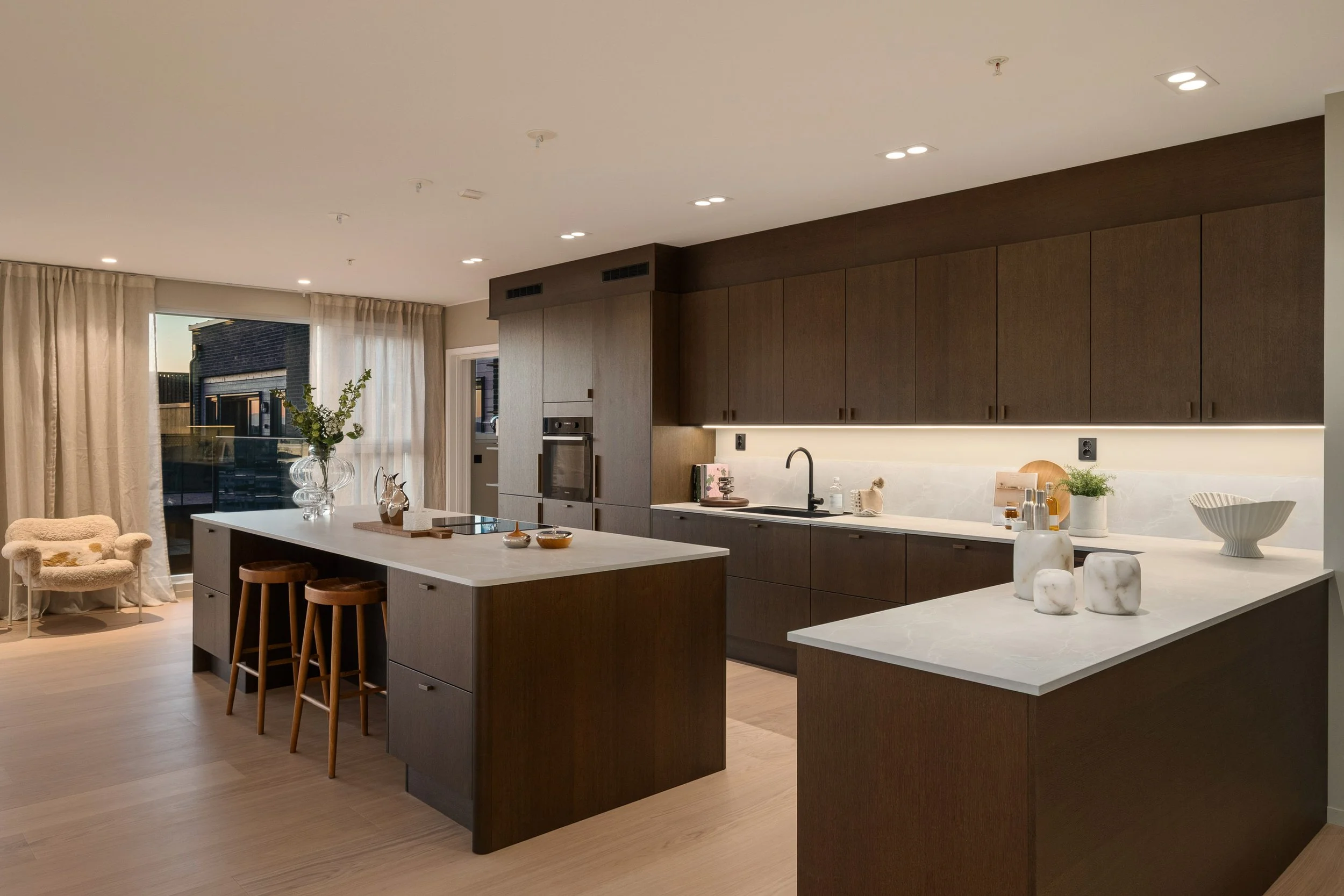 Moderne kjøkken med mørke tre- og hvite benkeplater, øy med trestoler, blomster og dekorative gjenstander, store vinduer med gardiner, og rolig belysning.