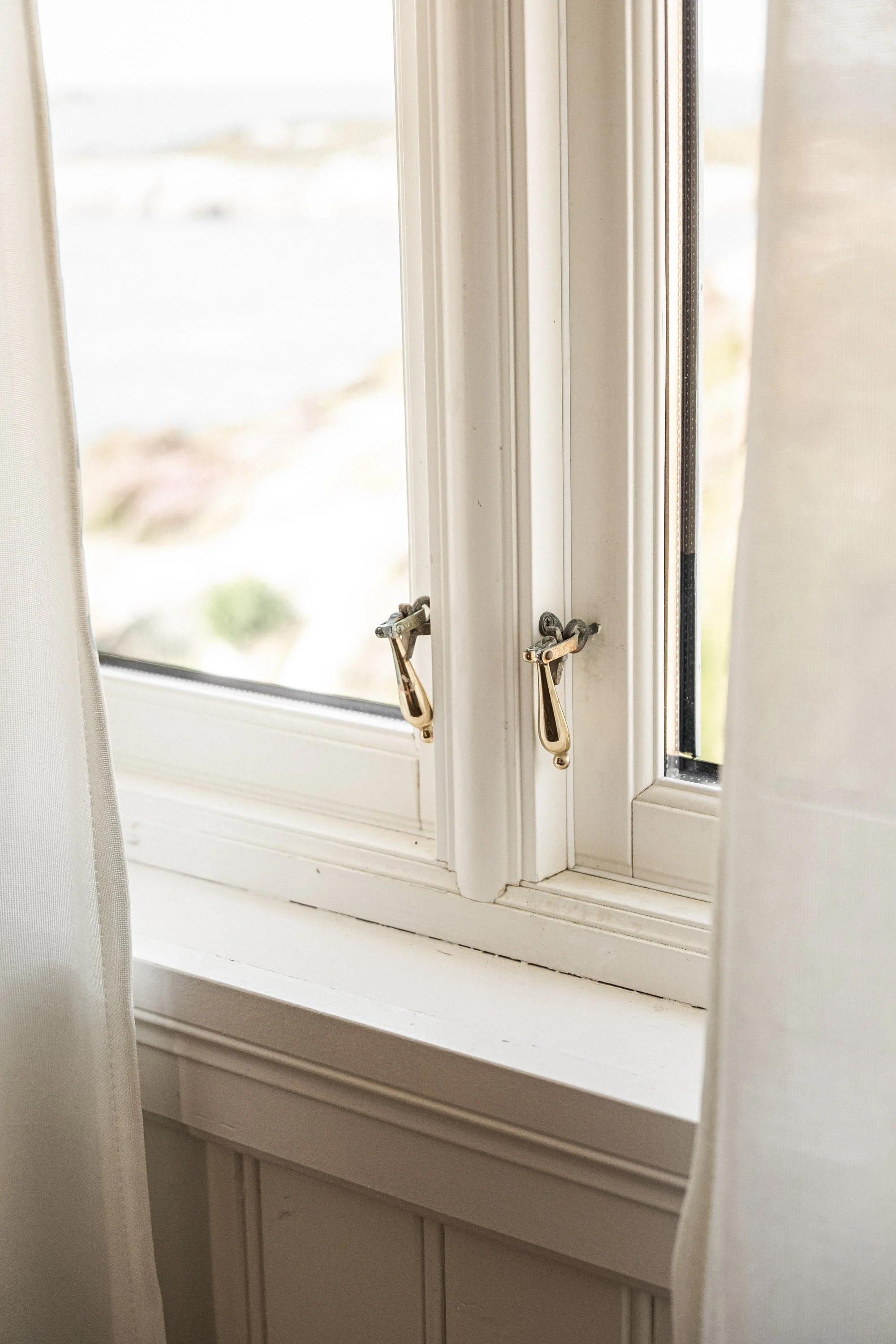 Nærbilde av en hvit vinduskarm med to vinduslåser, med gardiner på sidene.