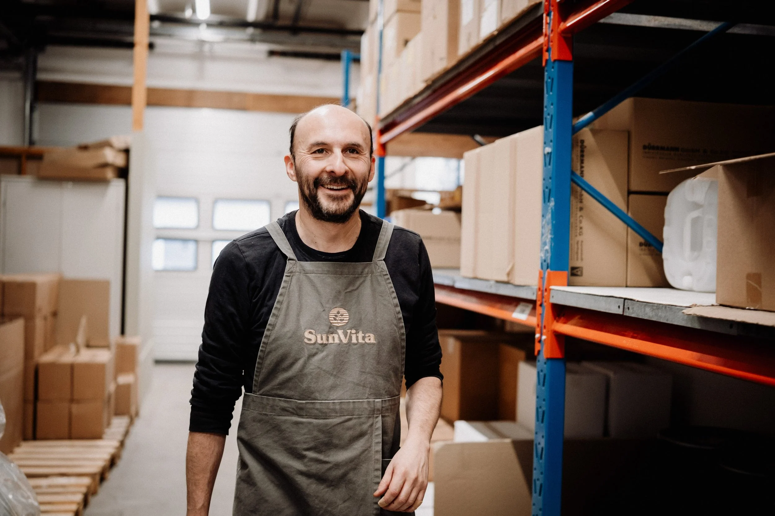 En mann smilende i et lager med hyller fulle av pappesker, iført forkle med logoen SunVita.