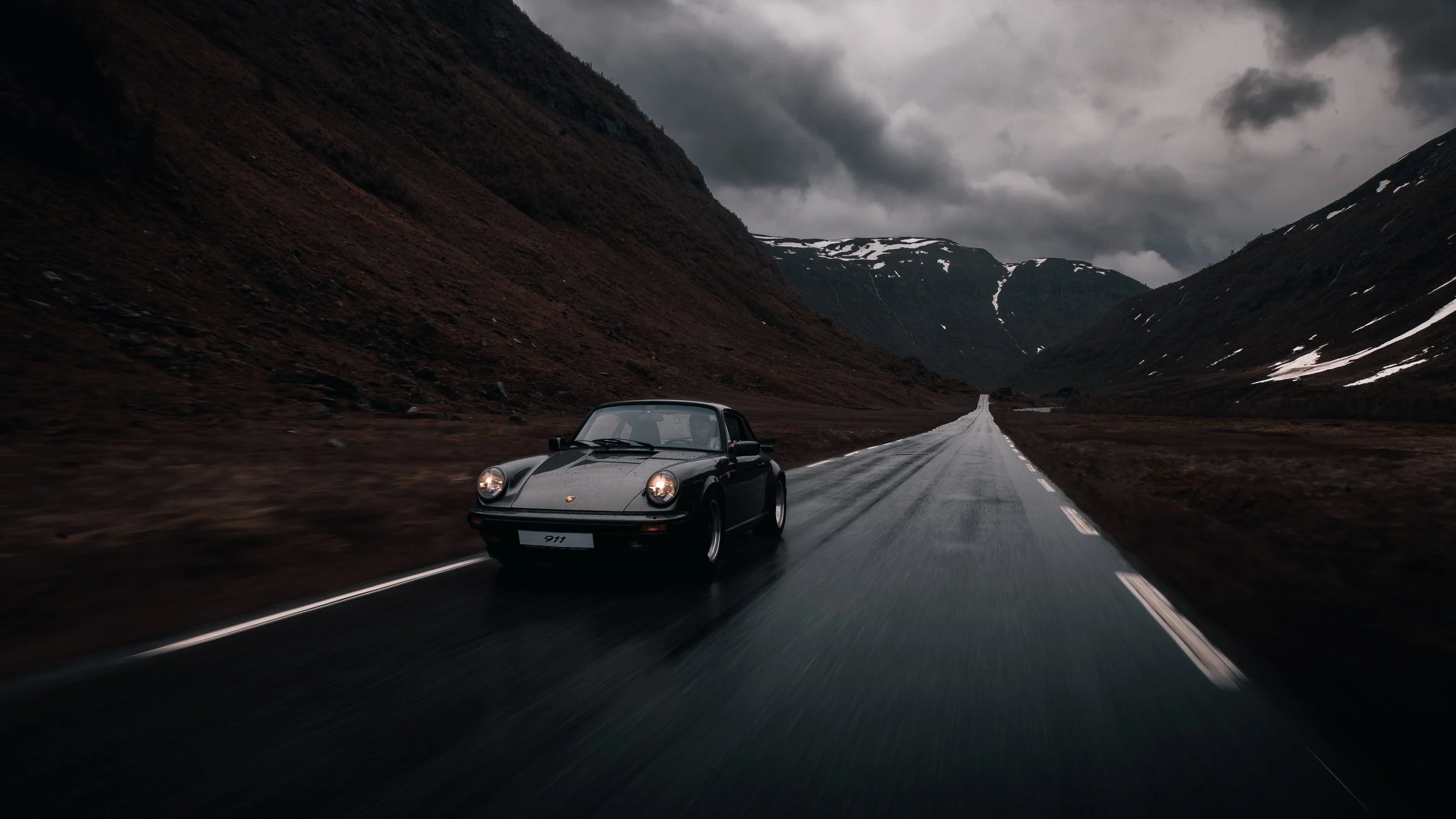 En svart Porsche 911 kjører på en tynn, våt asfaltvei gjennom fjellområder under en mørk, overskyet himmel.