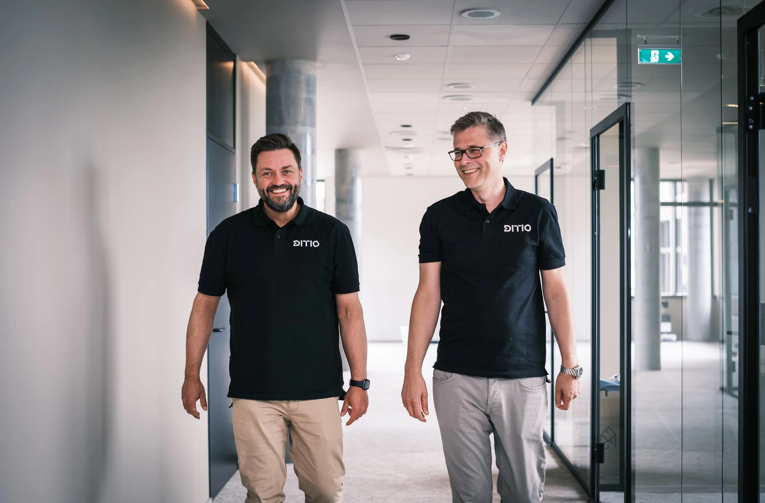 To menn går i en kontorbygning, smiler og har på seg svarte skjorter med selskapets logo, DITIO.