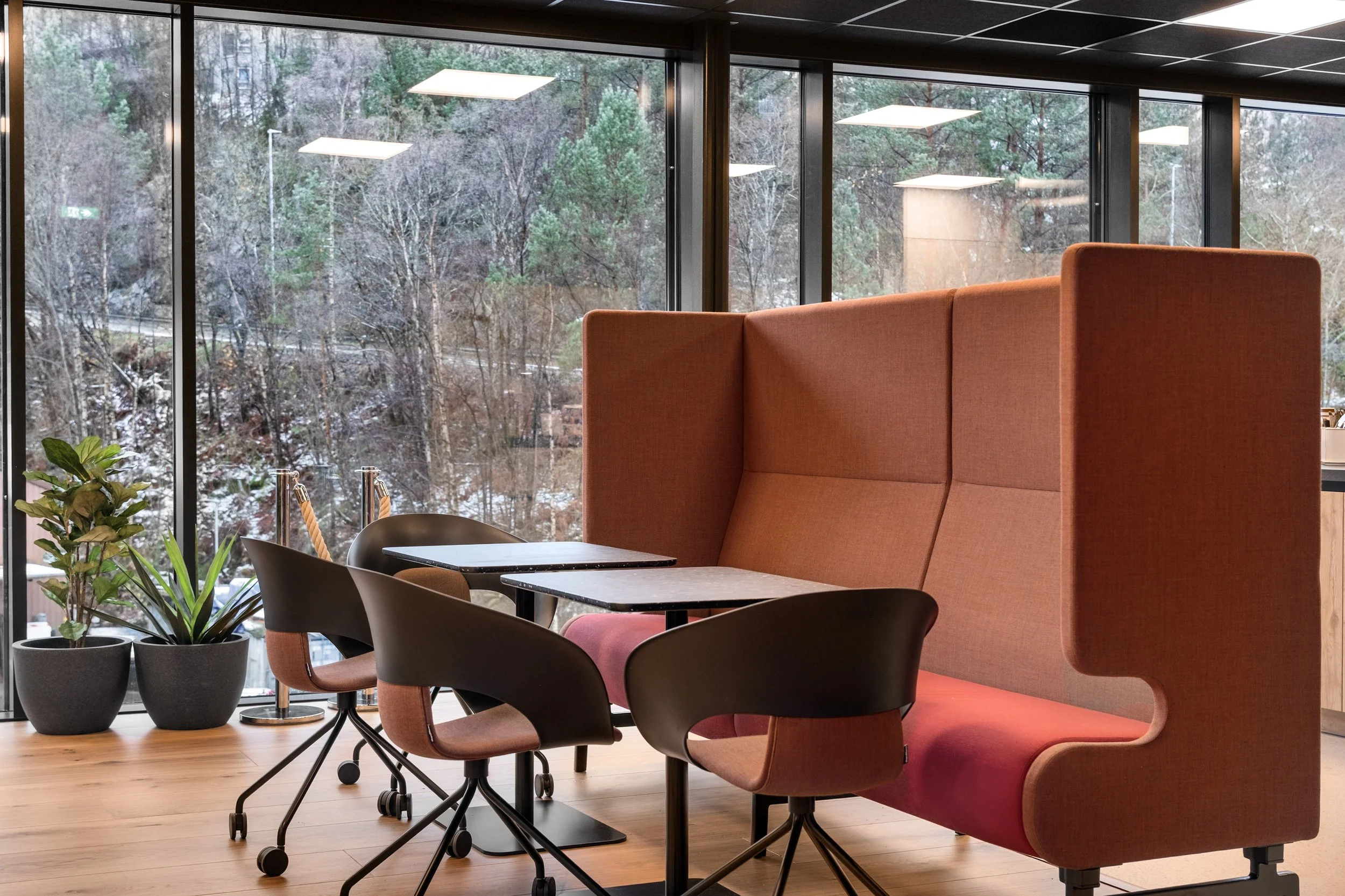 Moderne møtesone med store vinduer som viser utendørs natur, inkluderer høye planter, en høystolpe med papp og små bord med stoler.