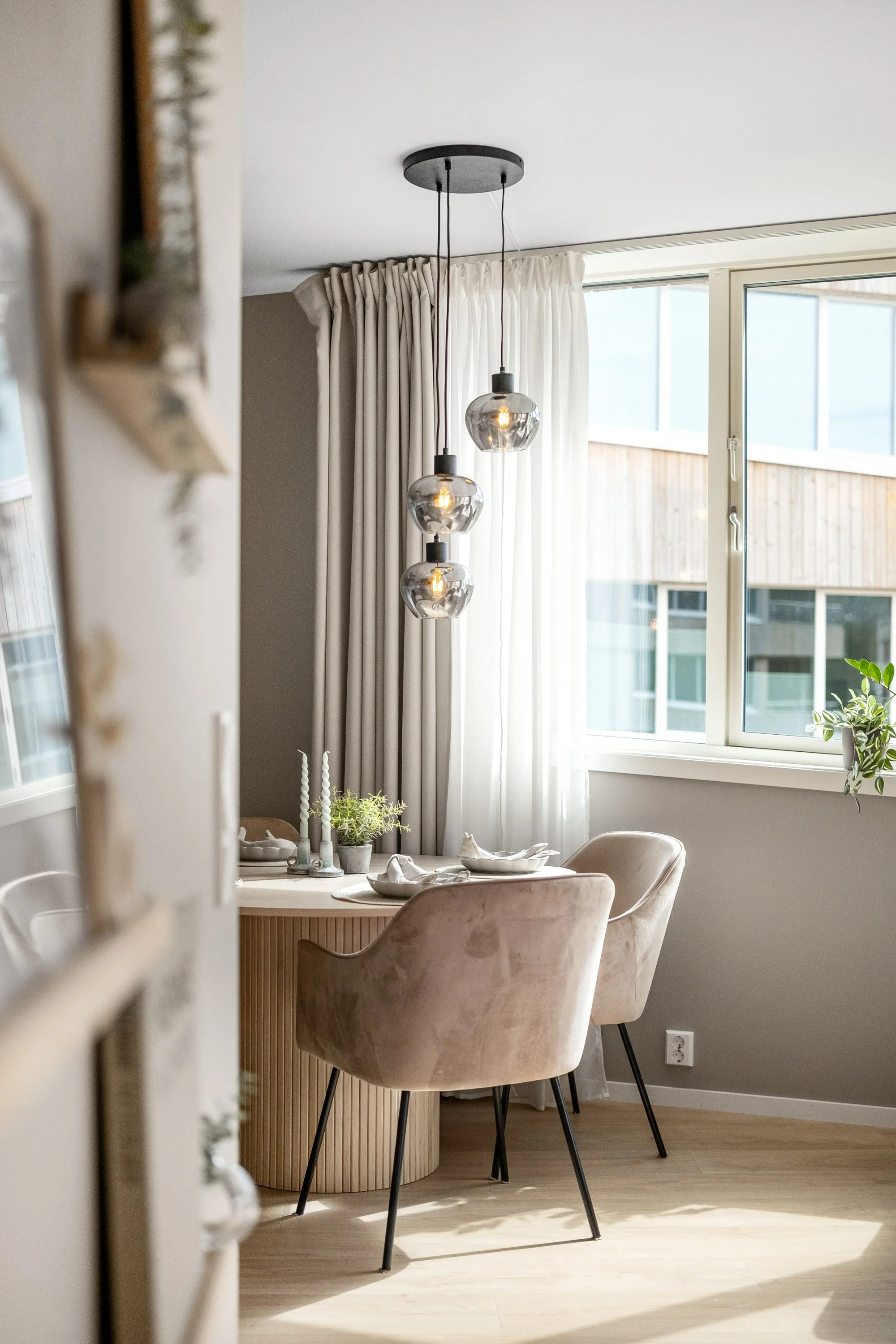 Et lyst spisestueområde med et rundt bord, to beige stoler med svarte ben, lys gardin og tre glasspendellamper over bordet. Naturlig lys slipper inn gjennom et vindu.