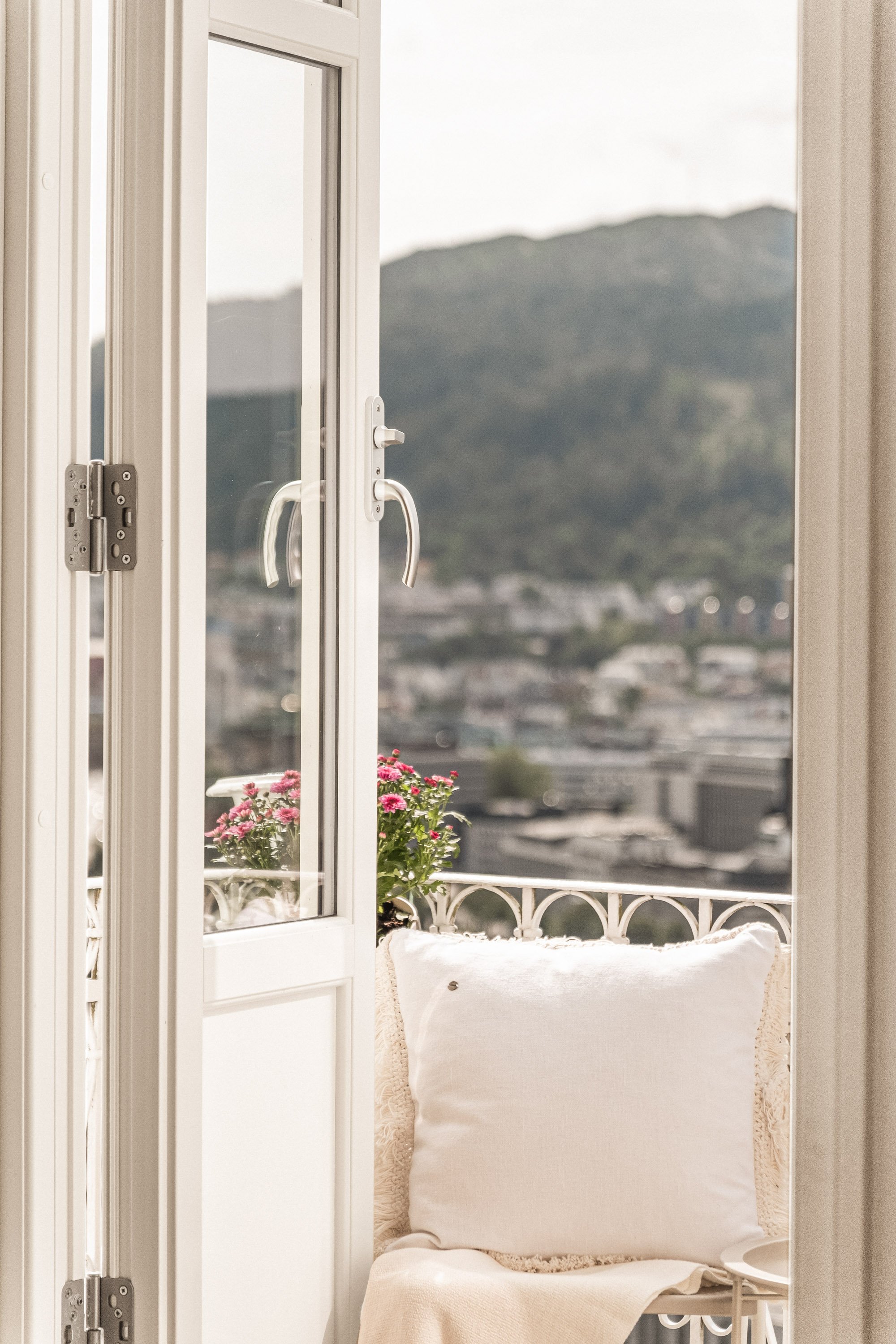 Utsikt fra en balkong med blomsterpotte og hvit pute, med fjell og by i bakgrunnen.