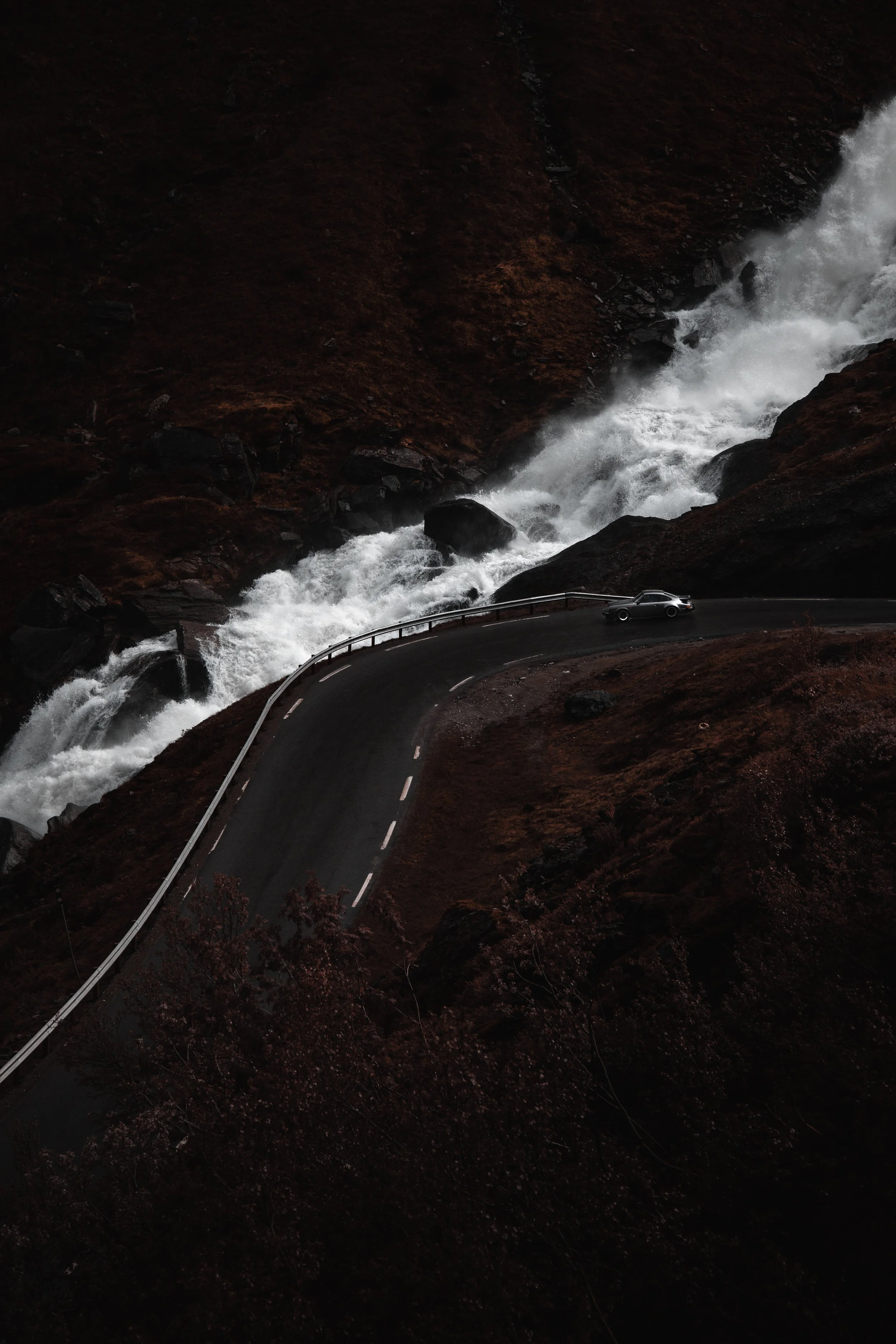 En svart bil kjører på en slyngende vei langs en foss med vann som fosser nedover klippene.
