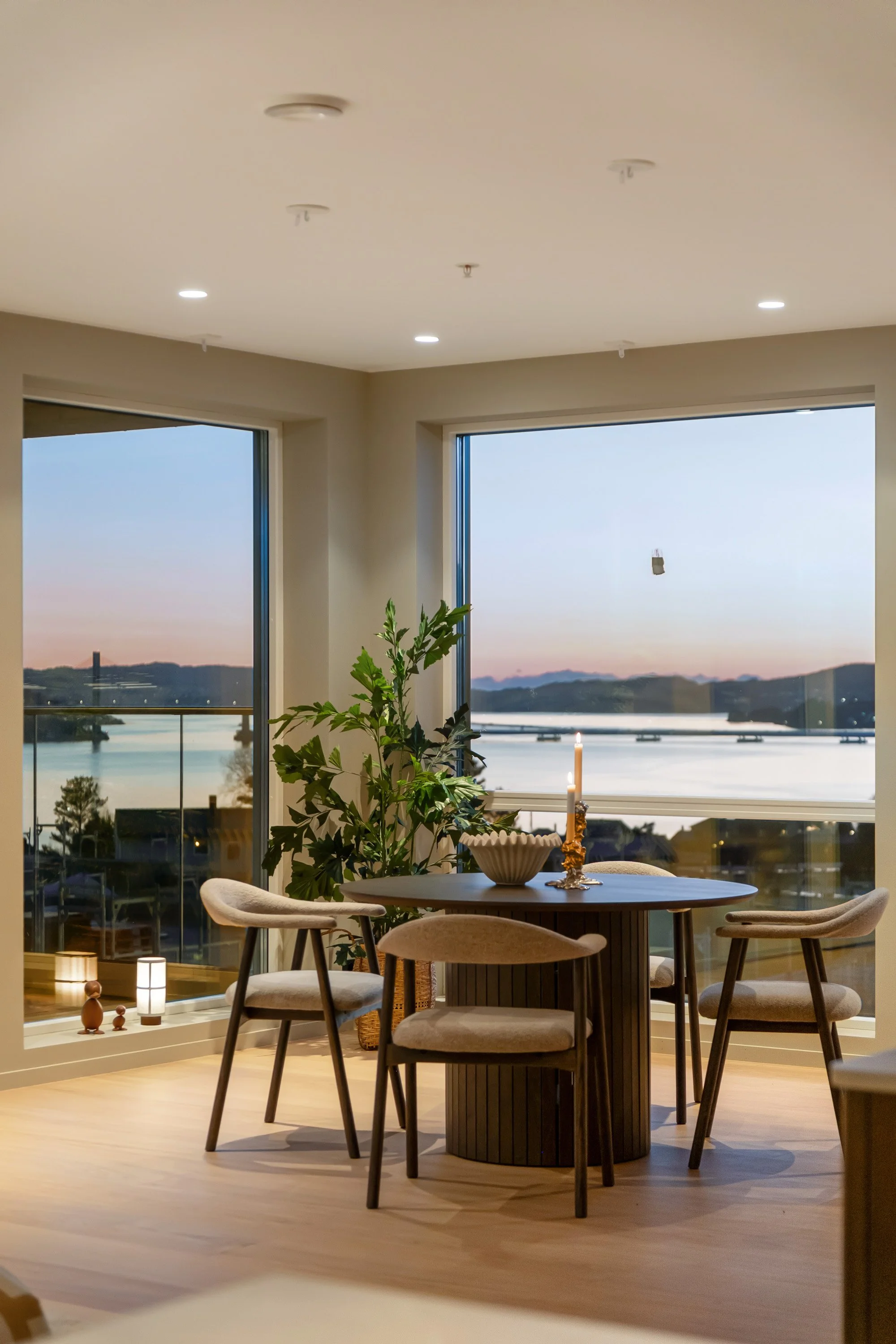 Innendørs stuebord med fire stoler, dekorativ lysestake, stor plante, store vinduer med utsikt over vann og fjell, solnedgang