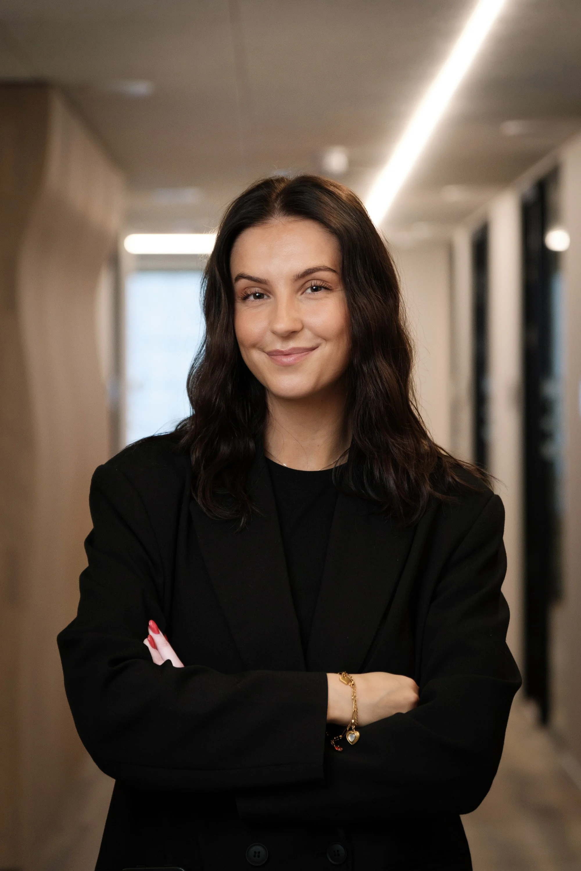 Young woman with dark brown wavy hair, wearing a black blazer, standing confidently with arms crossed in a modern indoor corridor.