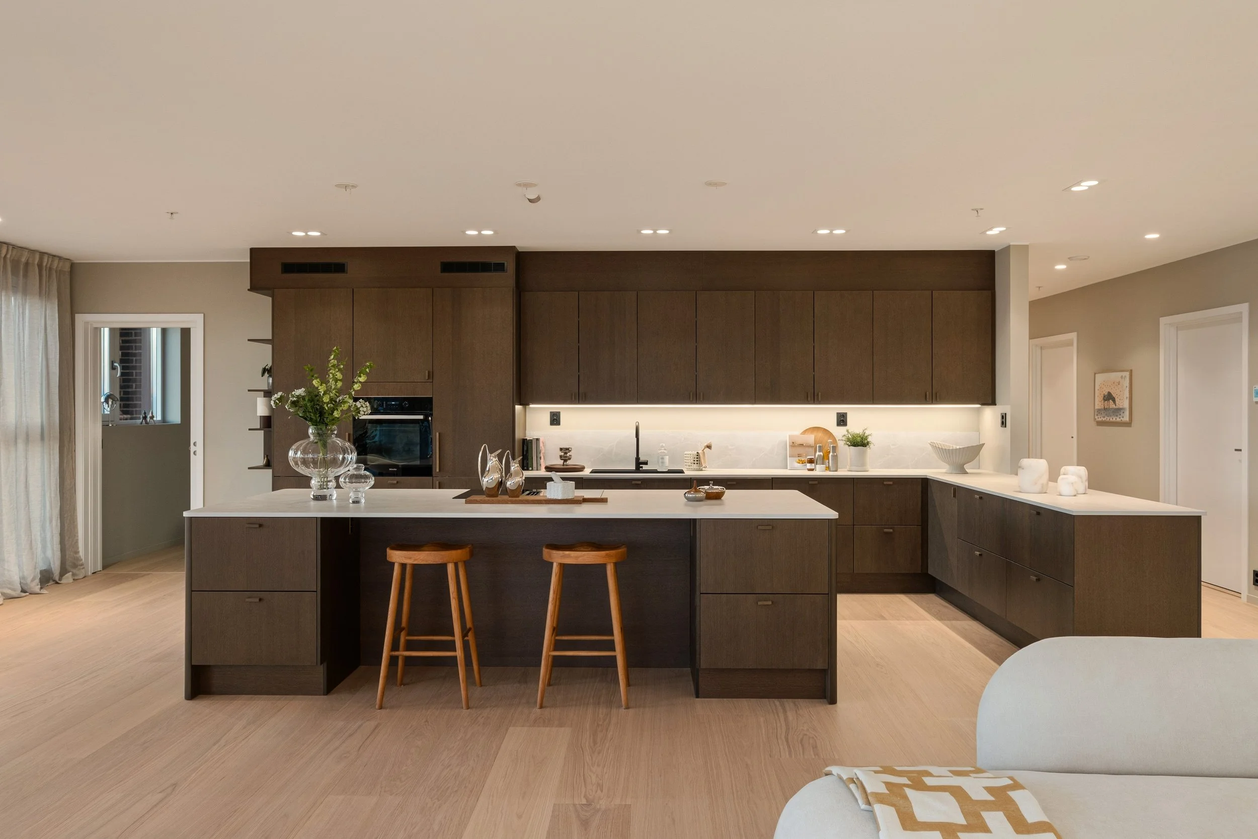 Moderne kjøkken med mørke trekjøkkenskap og lyse benkeplater, to trebarnstoler foran øyen, blomster i glassvase og dekorative elementer.