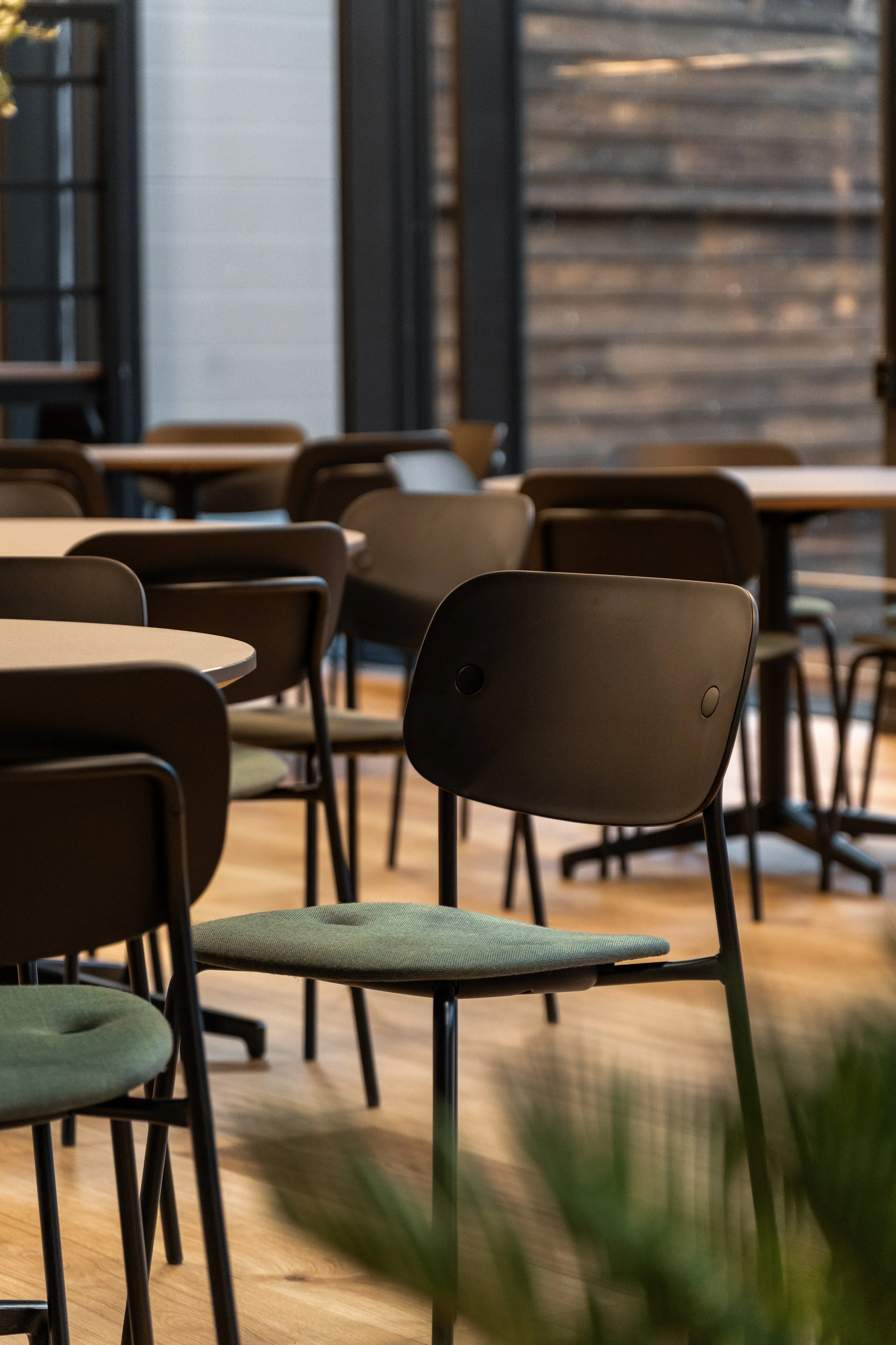 Innendørs terrasse med flere svarte stoler og bord, lyst tregulv, store vinduer og treverk på veggene.