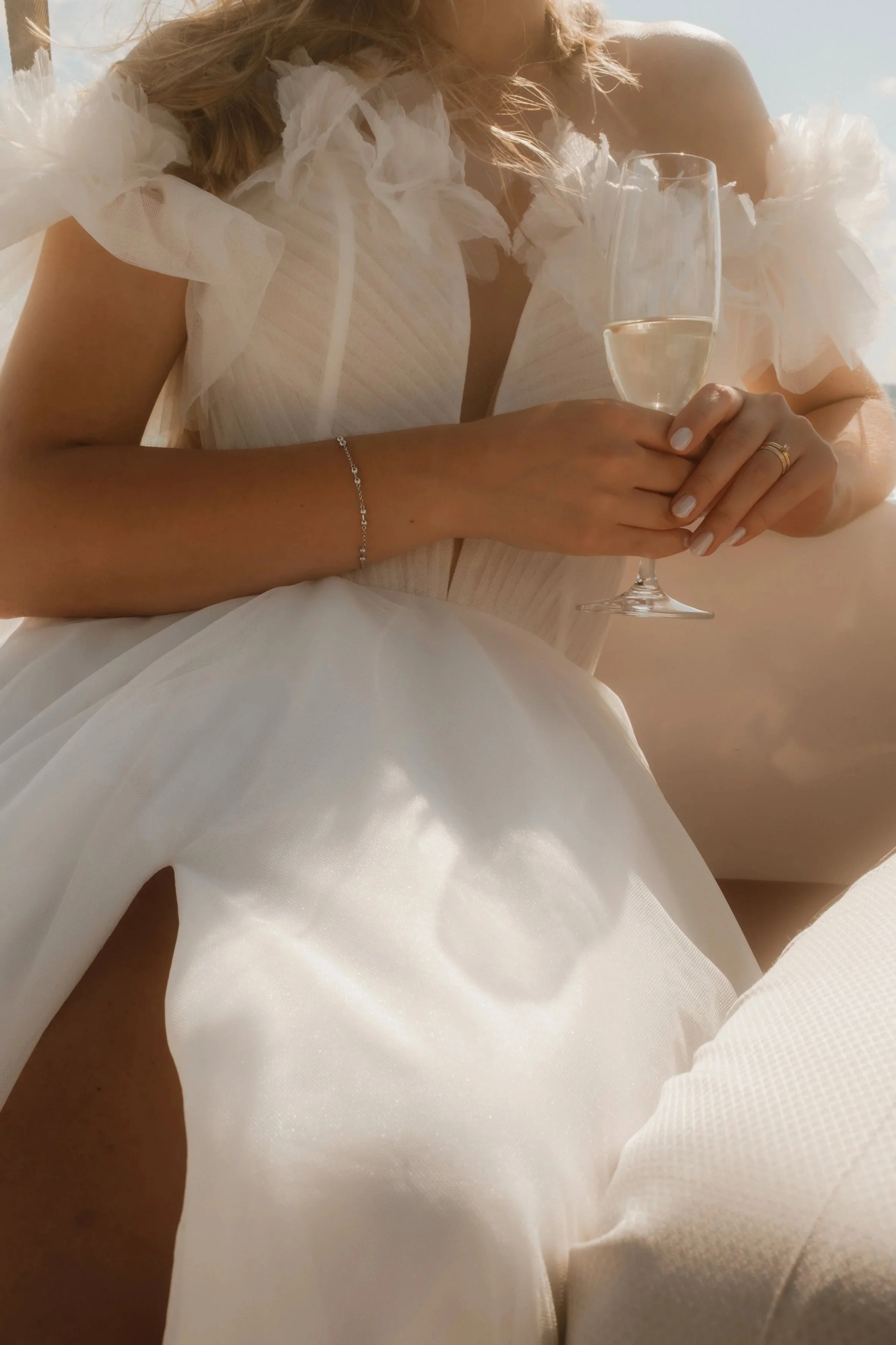 Eine Frau in einem weißen Kleid mit Tüll und Rüschen hält ein Glas Sekt. Sie trägt eine dünne Kette und einen Ehering. Das Bild ist in warmem Licht aufgenommen.