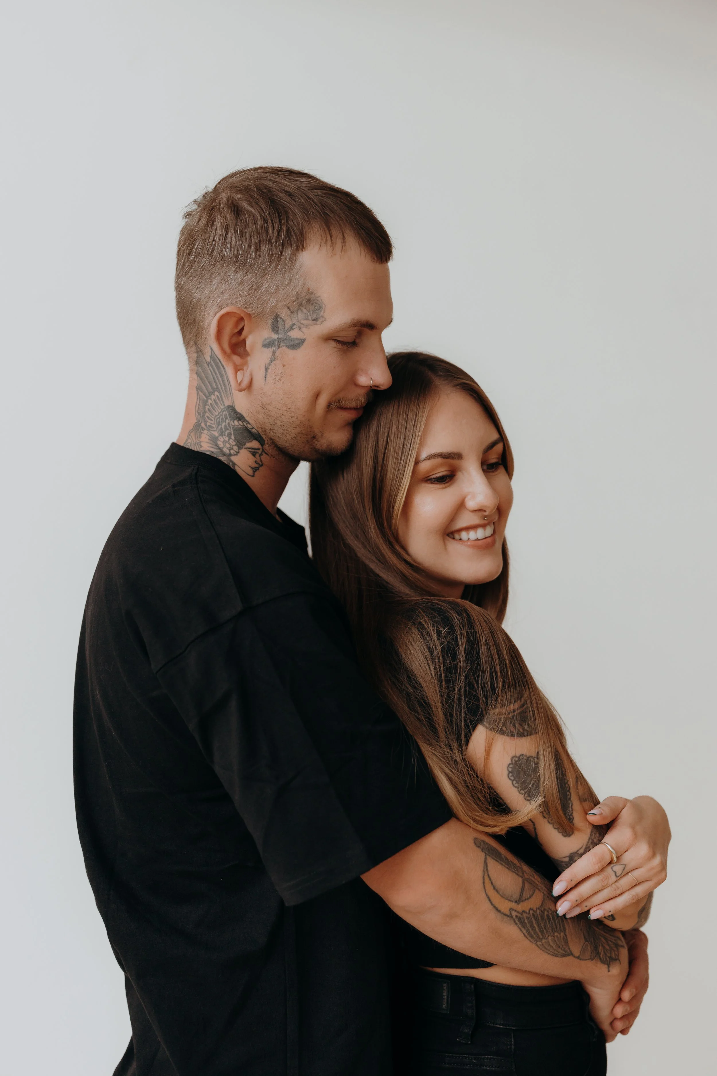 Ein Paar umarmt sich liebevoll vor einem weißen Hintergrund, beide haben Tattoos. Der Mann trägt ein schwarzes T-Shirt, die Frau trägt ein schwarzes Top.