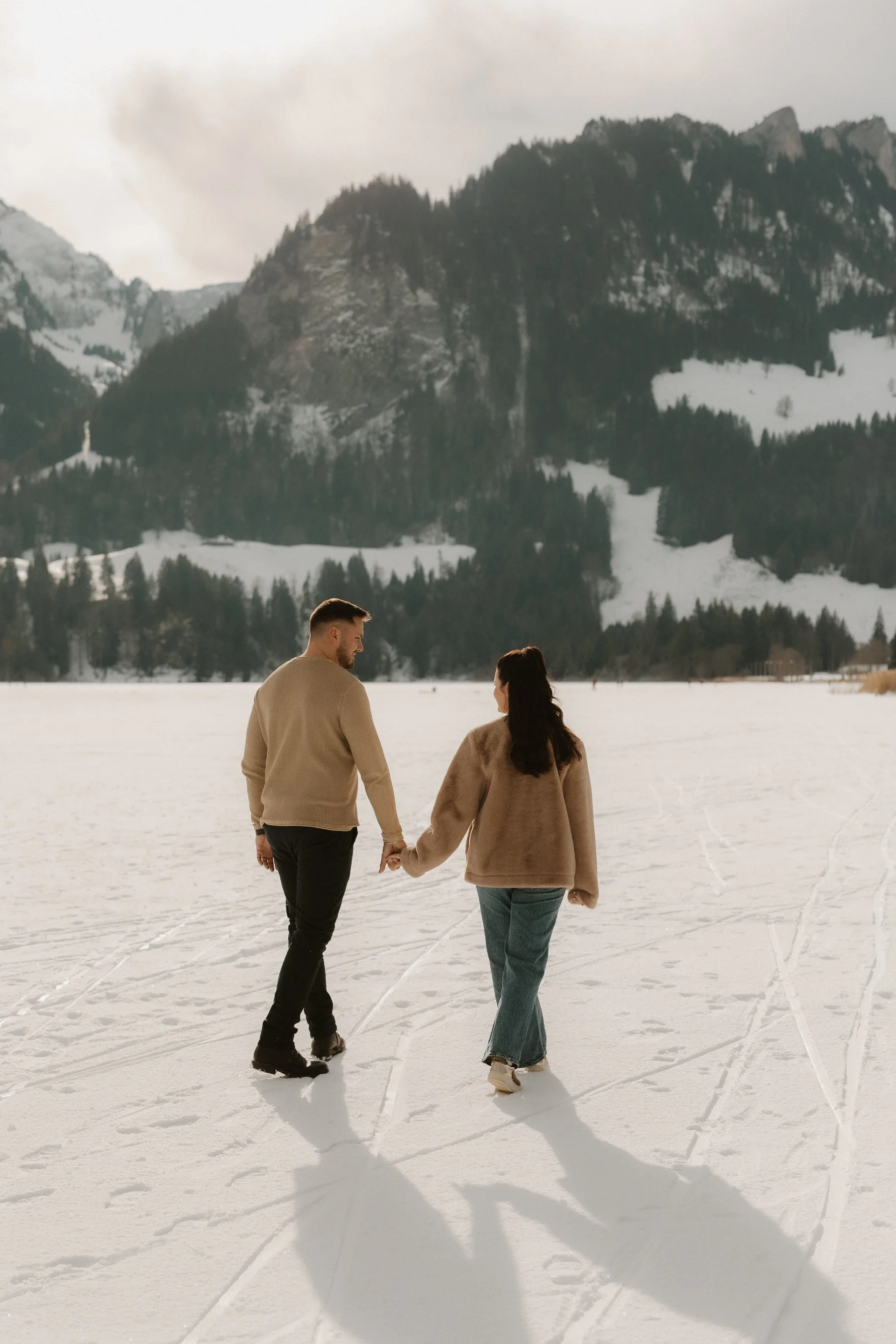 Ein Paar läuft Hand in Hand auf einem verschneiten See mit Bergen im Hintergrund, bei bewölktem Himmel.