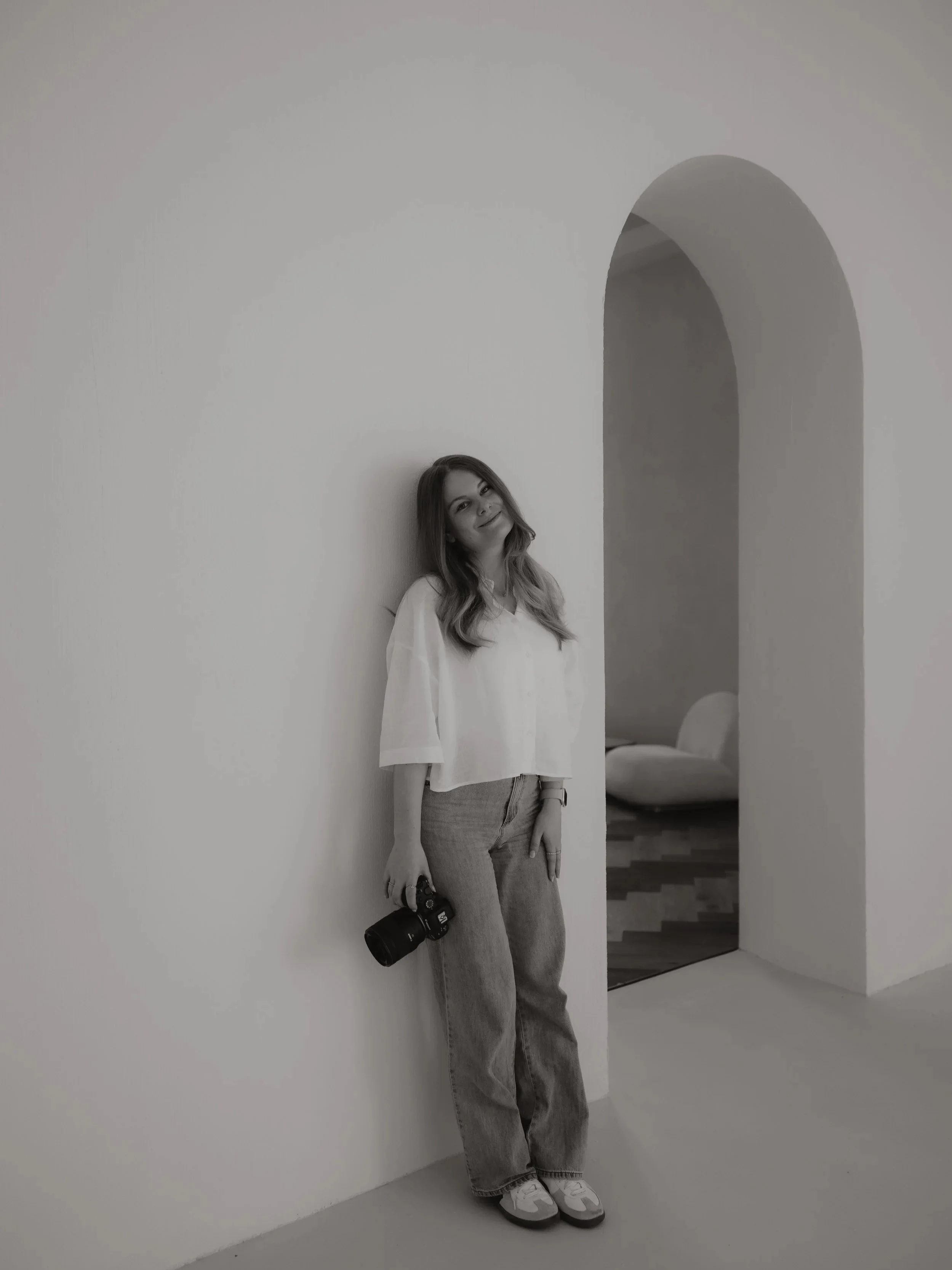 Junge Frau mit Kamera lehnt an einer weißen Wand in einem minimalistischen Raum.