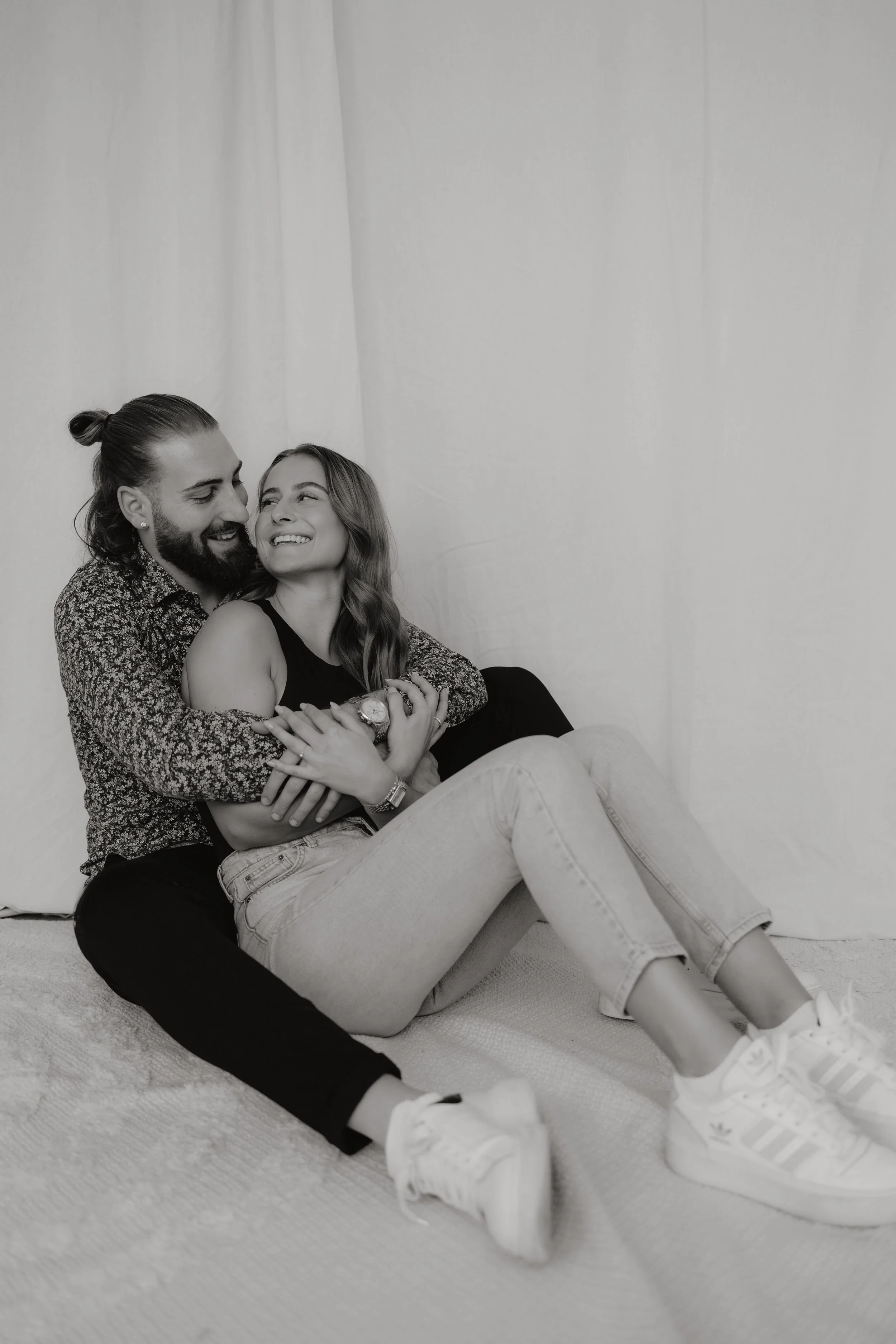 Ein lachendes Paar sitzt auf dem Boden, umarmt sich, vor einem weißen Hintergrund, schwarz-weißaufnahme.