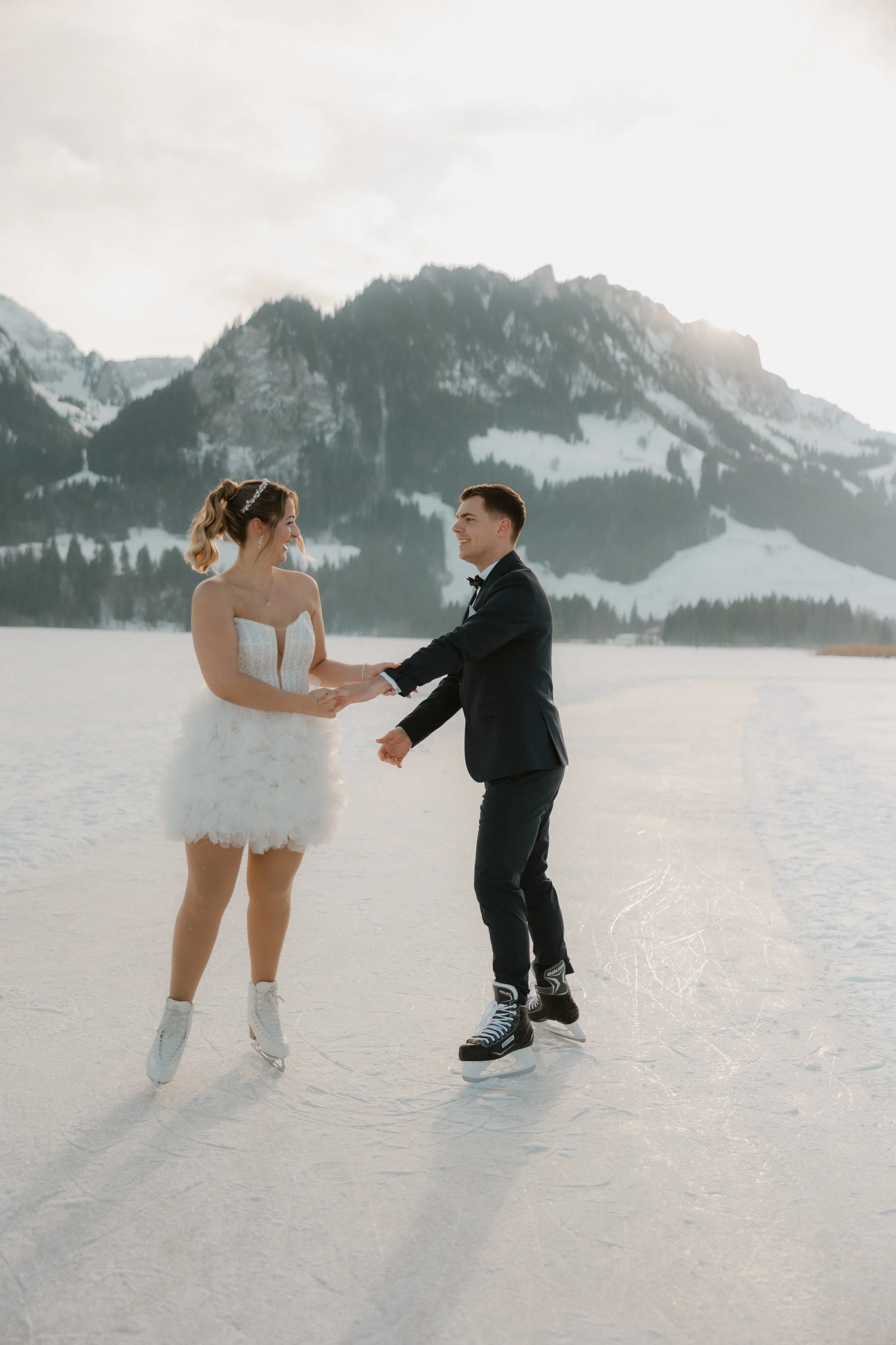 Ein Paar beim Eislaufen auf einem zugefrorenen See mit schneebedeckten Bergen im Hintergrund, das Hochzeitskleidung trägt.