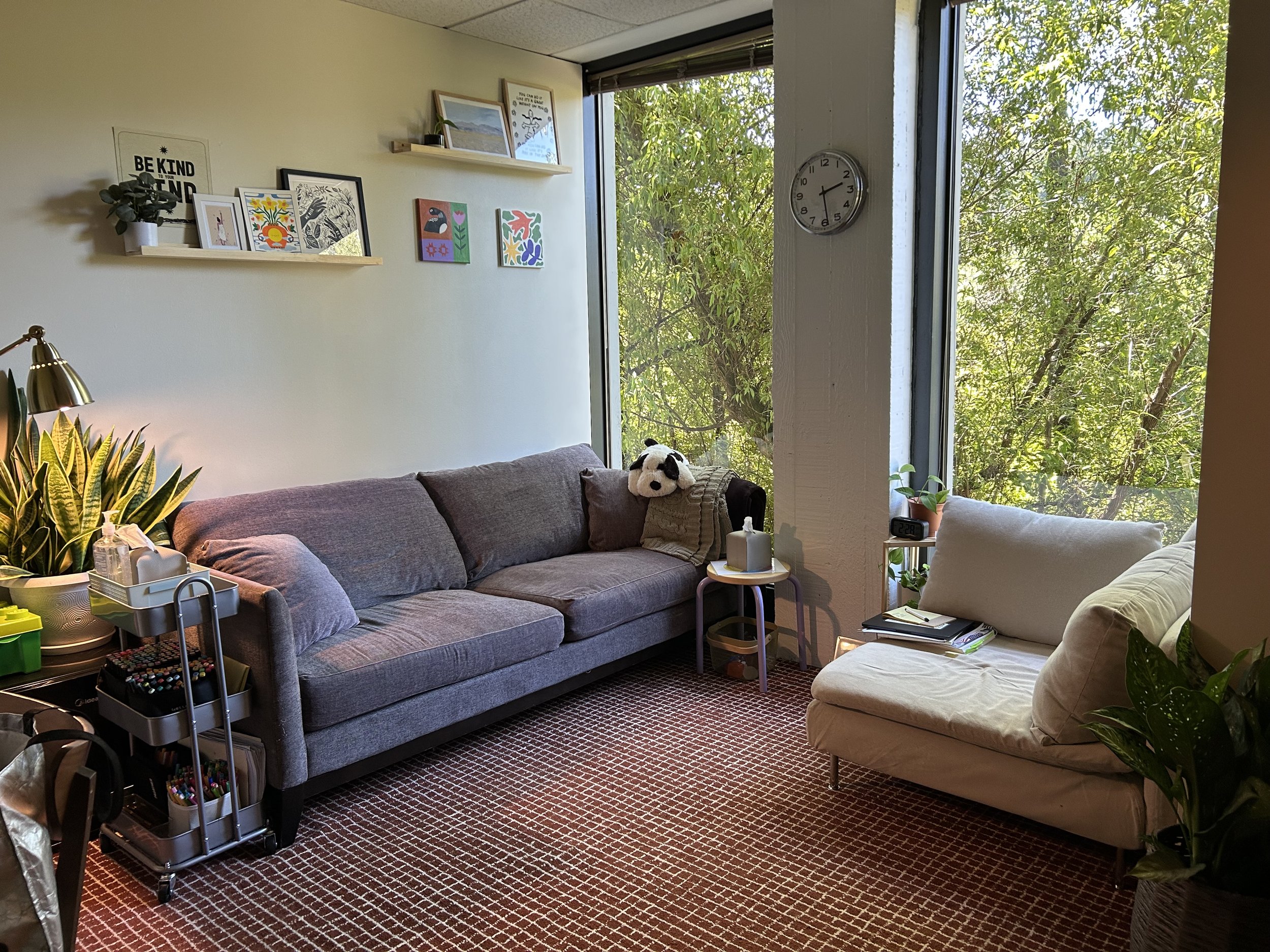 Living room with a gray sofa, white armchair, large window with view of trees, wall clock, wall art, houseplants, side tables, and a red patterned carpet.