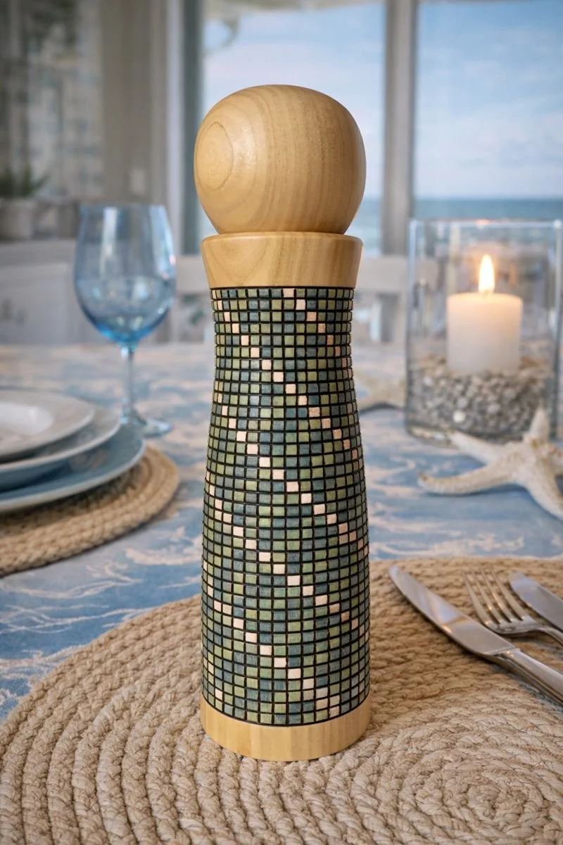 Decorative salt or pepper shaker with a mosaic pattern on a dining table near a lit candle and dishware, with a view of the ocean through large windows.