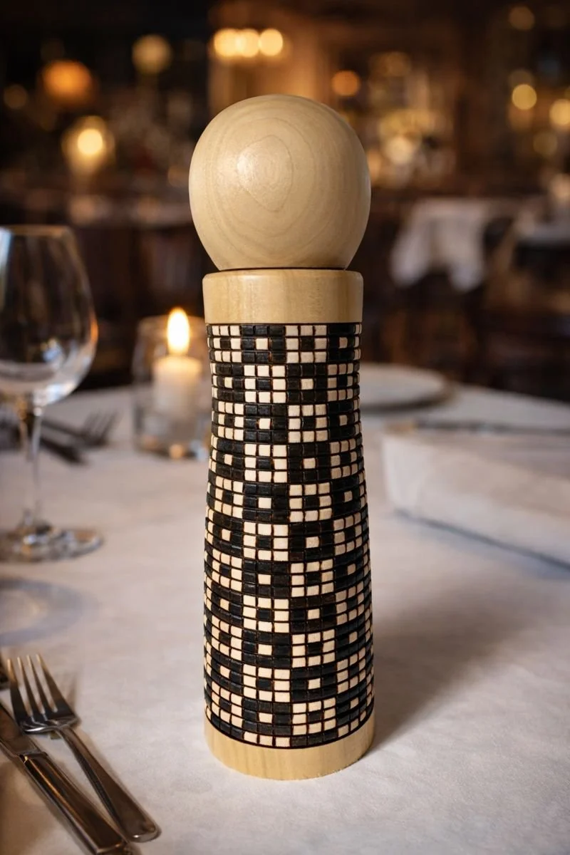 A wooden pepper grinder with a decorative black and white geometric pattern, placed on a table in a dimly lit restaurant setting.