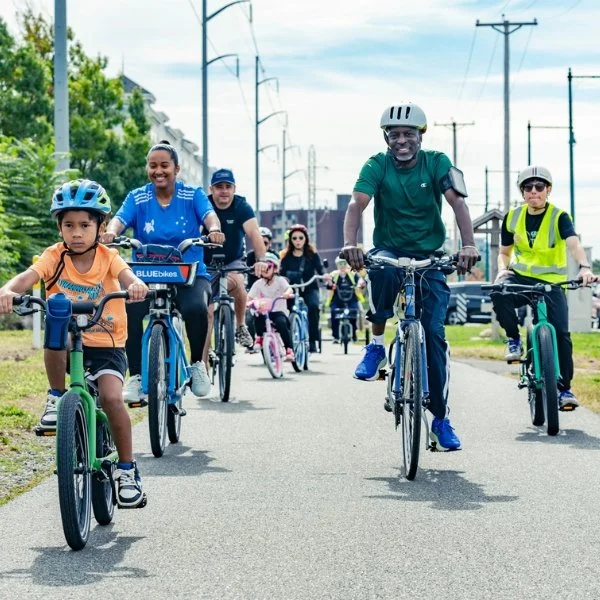A group of cyclists of different ages, ethnicities, and ability levels riding down a rail trail on a sunny day.