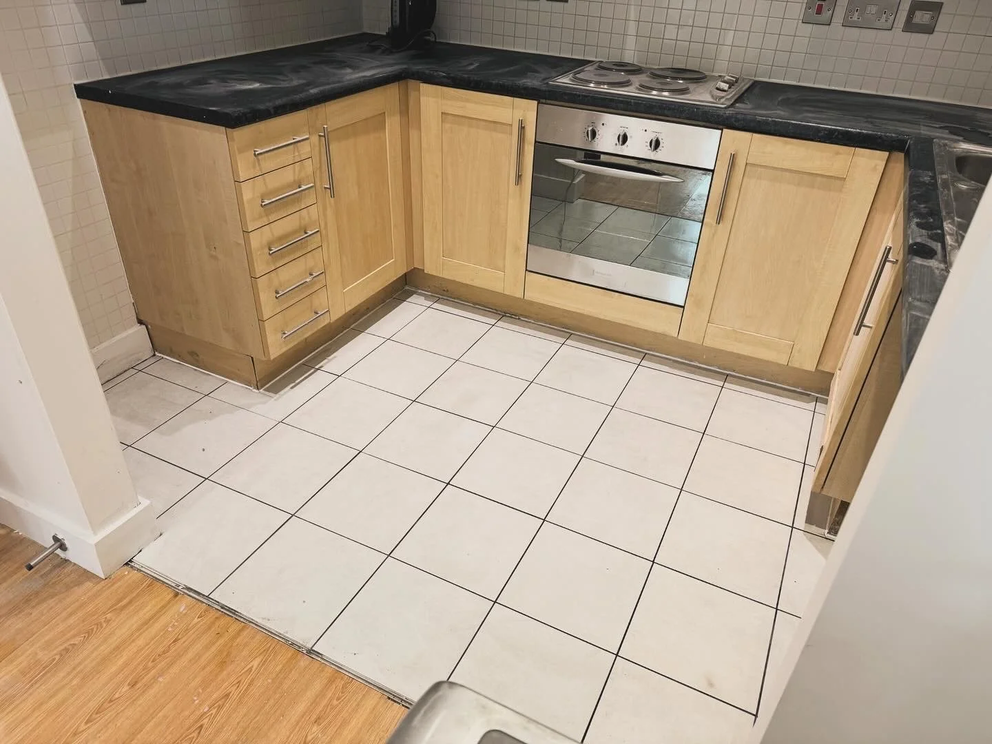 Kitchen with wooden cabinets, a black countertop, and a stainless steel oven in a corner. The floor has white tiles, with some needing replacement or repair, especially near the adjacent wooden floor.