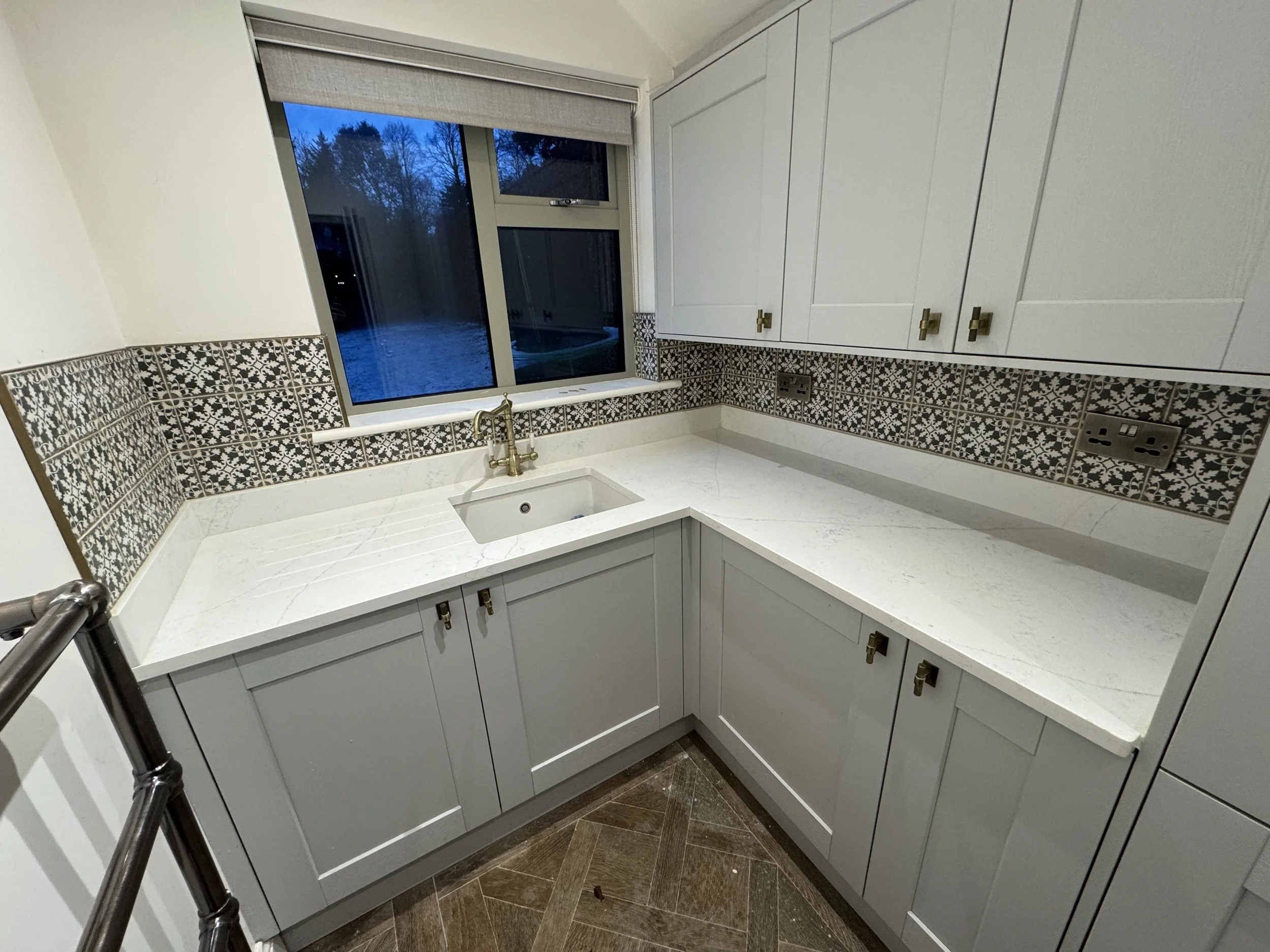 A kitchen corner with white cabinets, a white marble countertop, a small under-mount sink with brass faucet, decorative black and white tile backsplash, and a window showing an outdoor scene with trees and a blue sky.