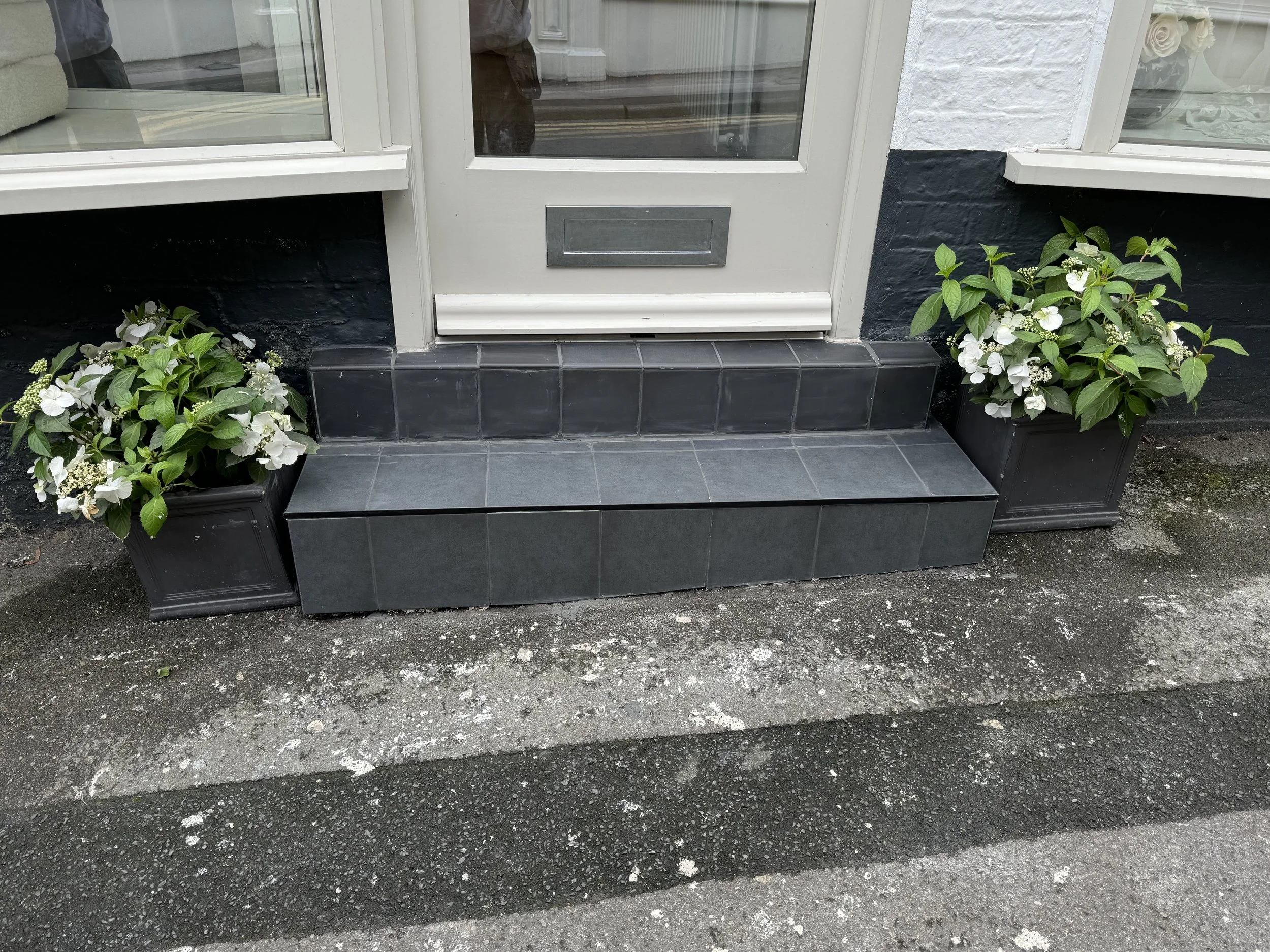 Black tiled steps leading to front door with white flowers in black planters on either side.