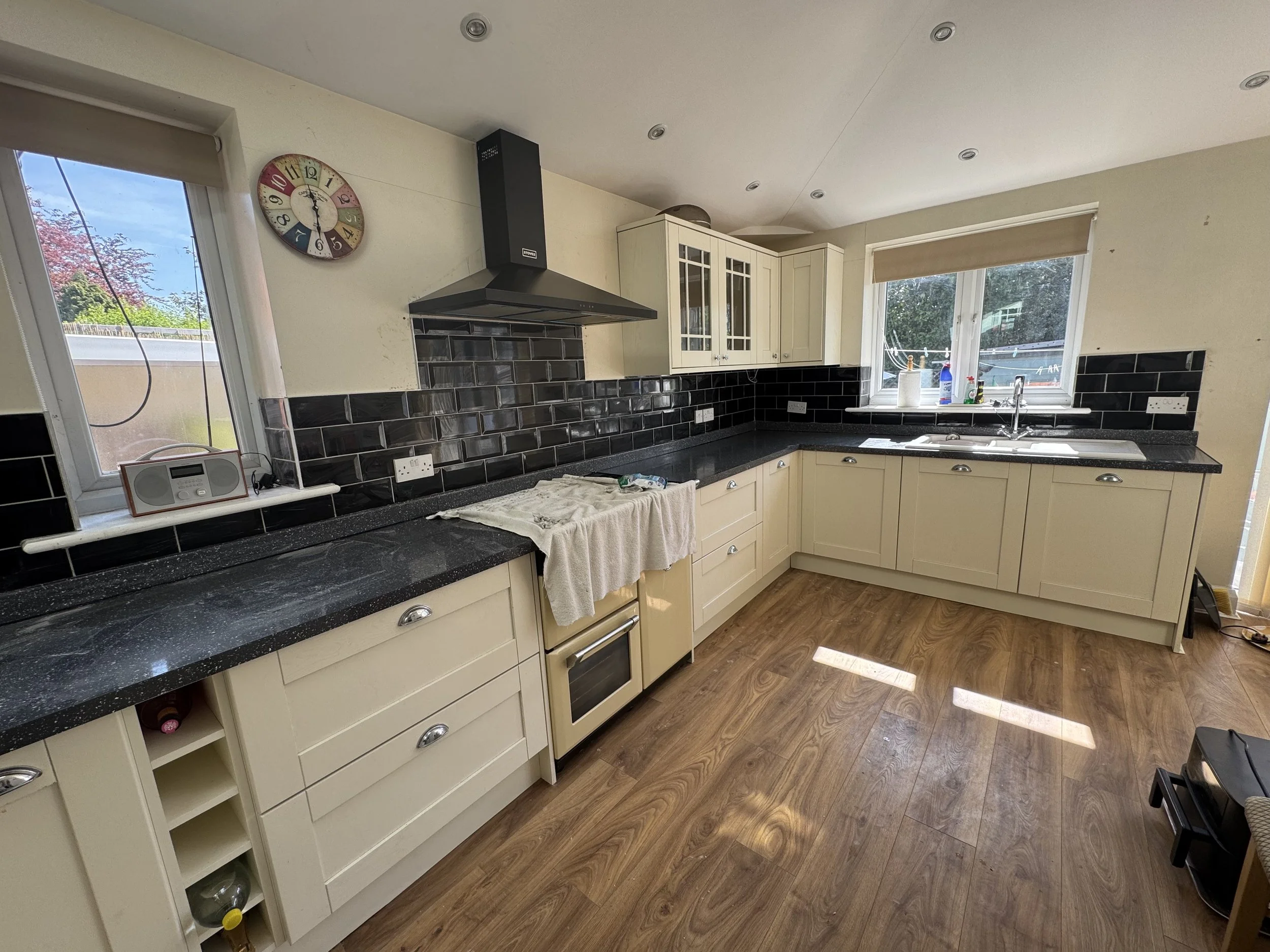 Kitchen with cream-colored cabinets, black countertop, black tile backsplash, a window over the sink, and a wall clock.