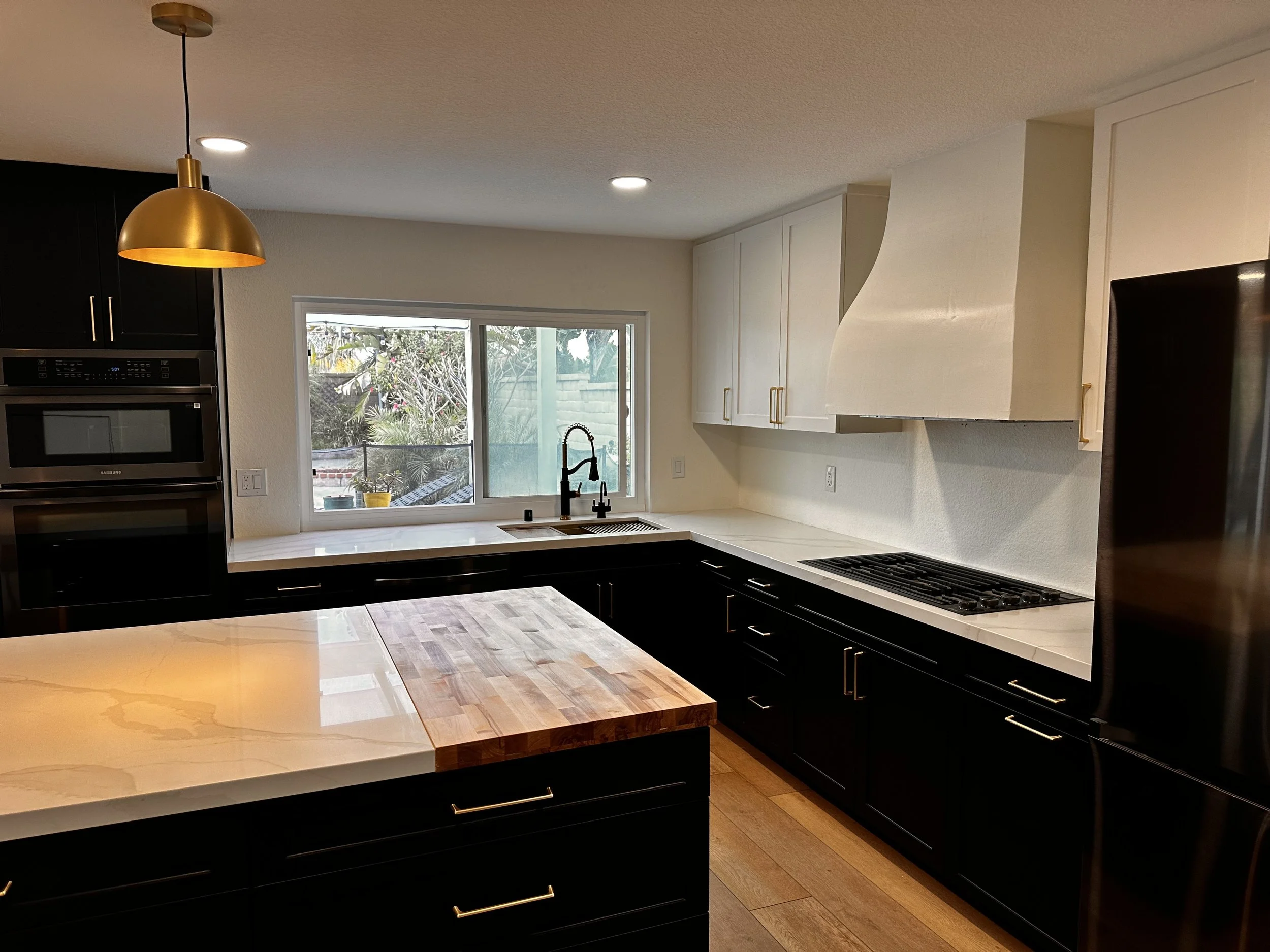 Modern kitchen with white cabinets, black lower cabinets, wooden and marble countertops, stainless steel oven, black faucet, large window overlooking garden, and pendant light above island.