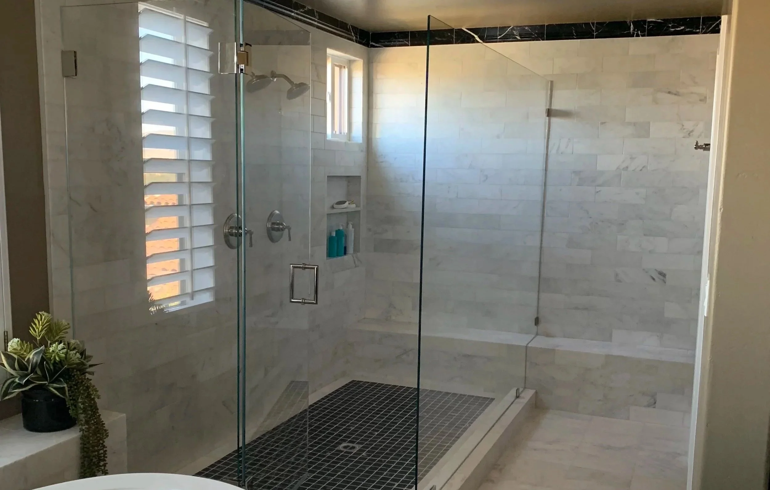 Modern bathroom with walk-in shower featuring glass walls, black tile floor, and white brick-style tile walls, with a window and plants outside the shower.