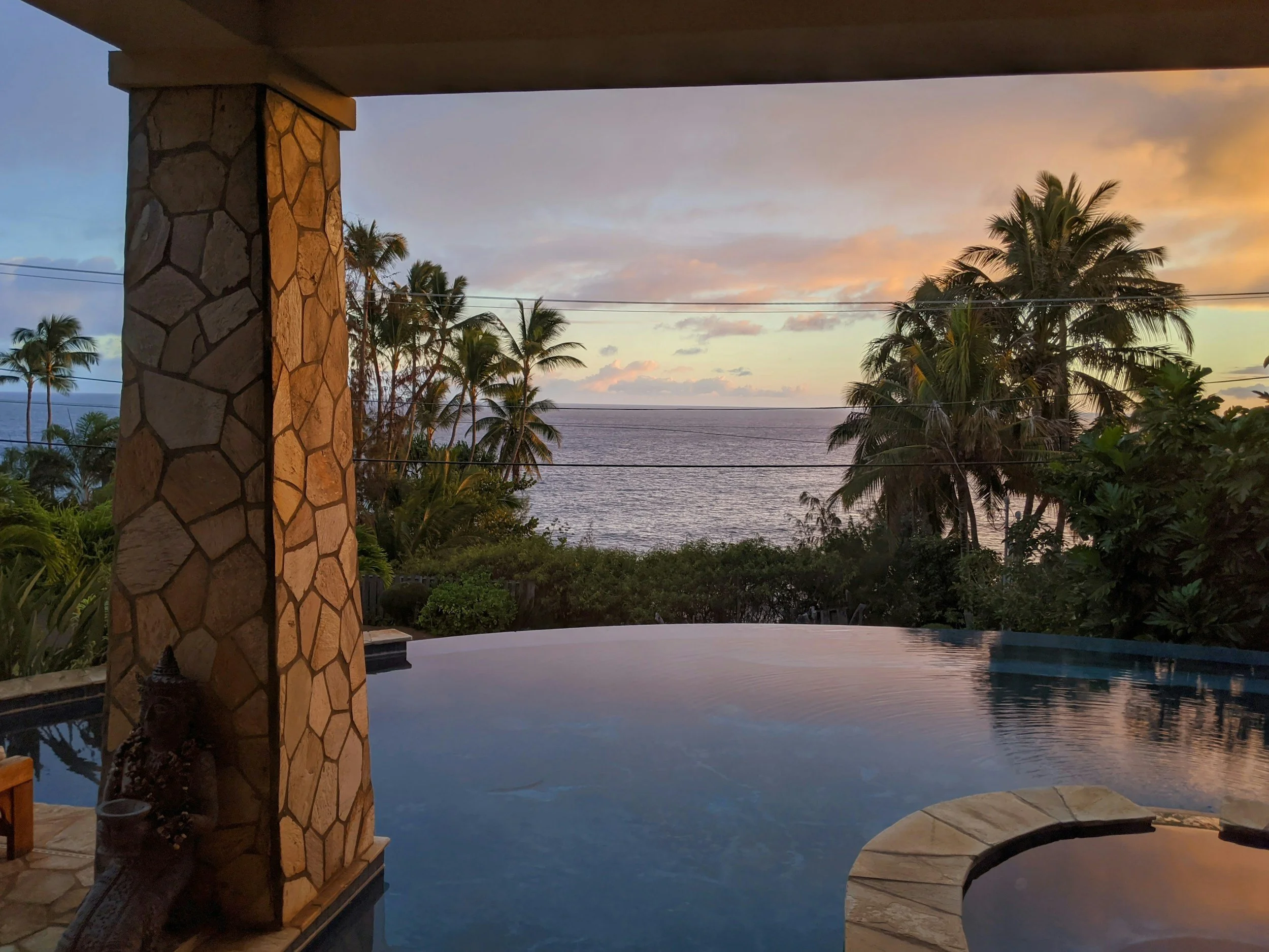 View of a sunset over the ocean with palm trees, seen from a patio area with a stone pillar and a swimming pool in the foreground.
