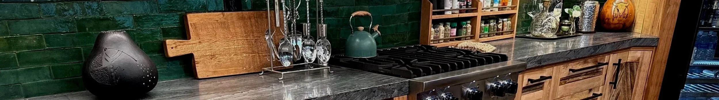 Kitchen countertop with various items including a black decorative vase, a wooden cutting board, a utensil holder with spoons, a green teapot, spice jars, glass vases, and a stack of books.