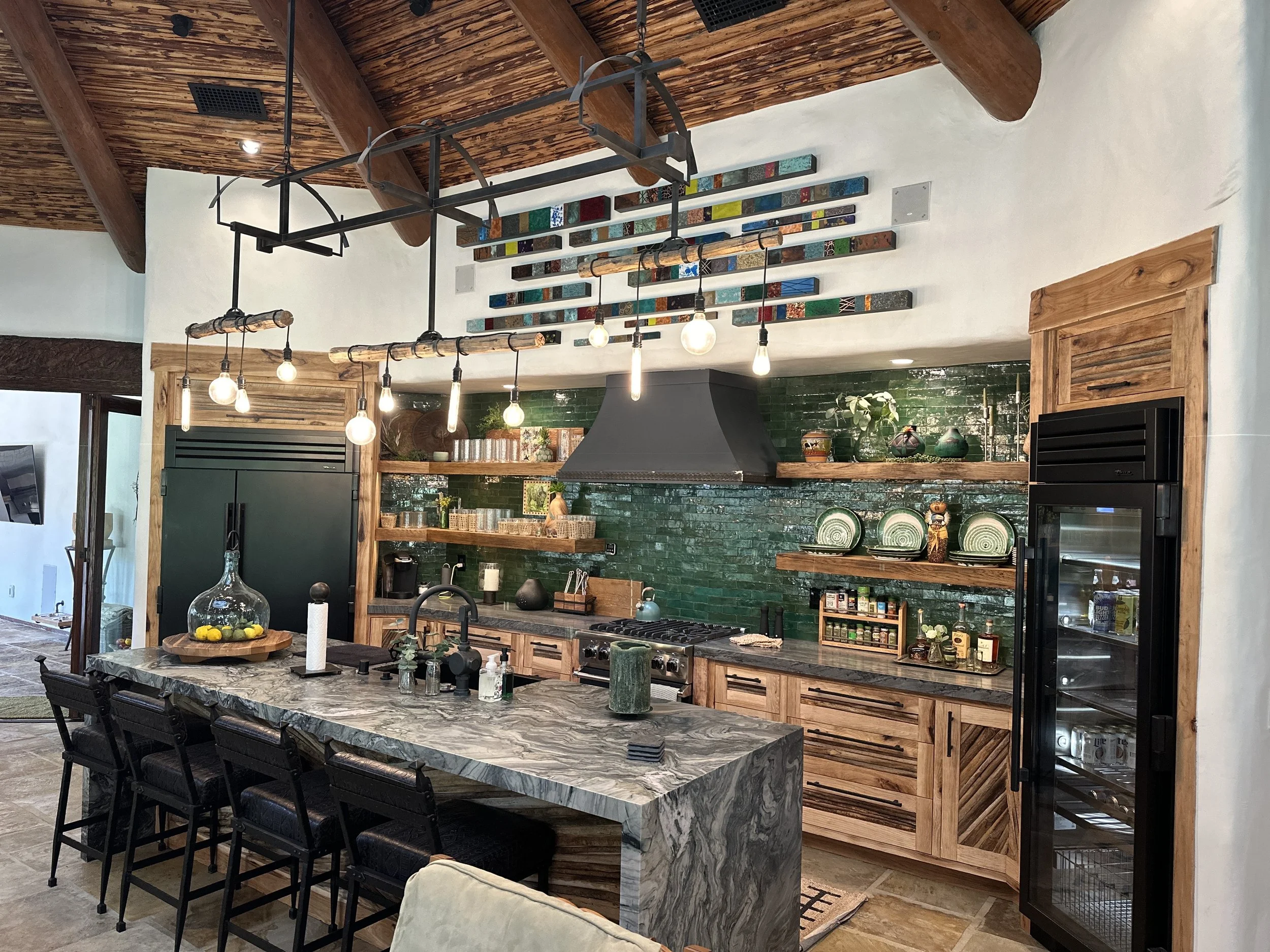 A Spanish modern kitchen with a high wooden, vaulted ceiling, a black marble kitchen island with black chairs, and a green tiled backsplash. Decor includes pots, vases, and tableware, with lighting fixtures hanging above the island.