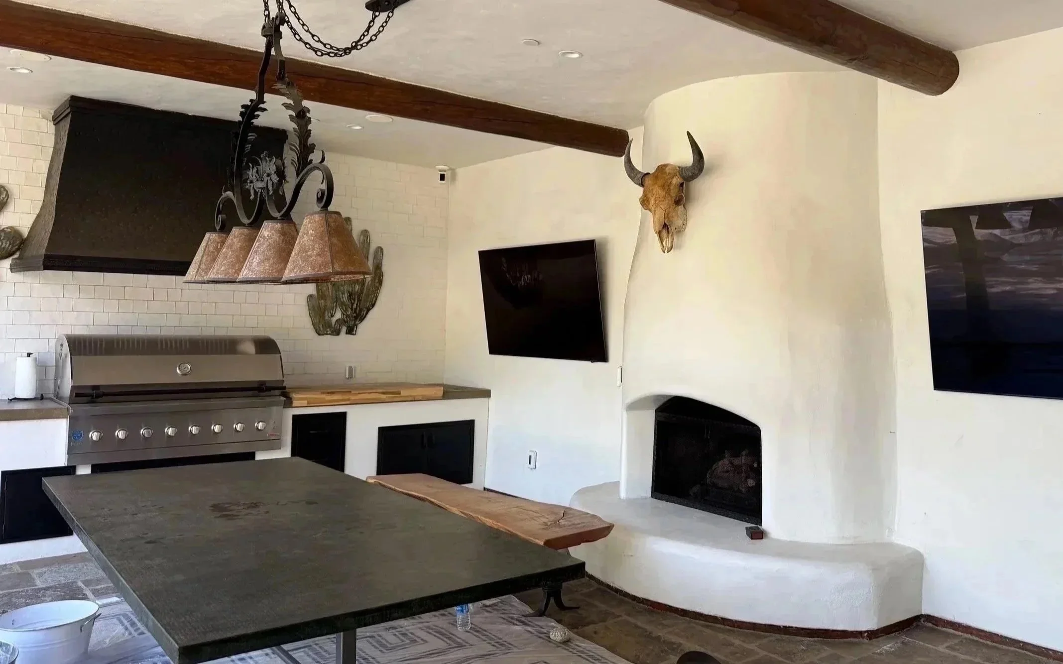 The image shows a kitchen and living room area with a large stone island in the foreground, a built-in grill, a hanging chandelier, and a television mounted on the wall. There is a decorative animal skull with horns mounted on the wall.