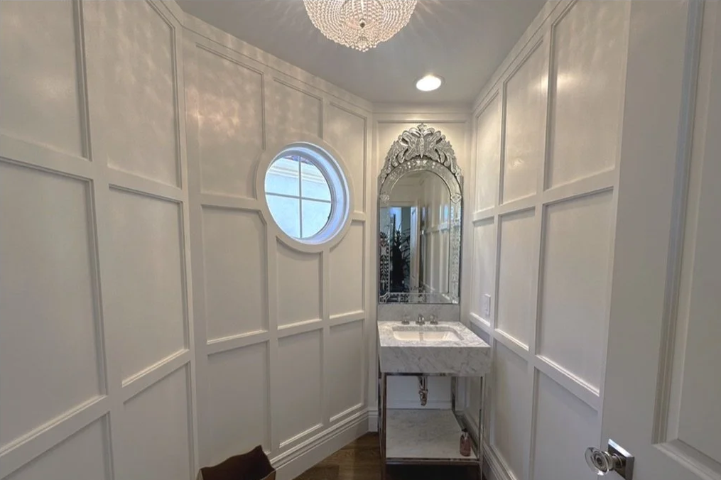 Small bathroom with white paneled walls, a round window, a marble vanity with a decorative mirror, and a chandelier ceiling light.