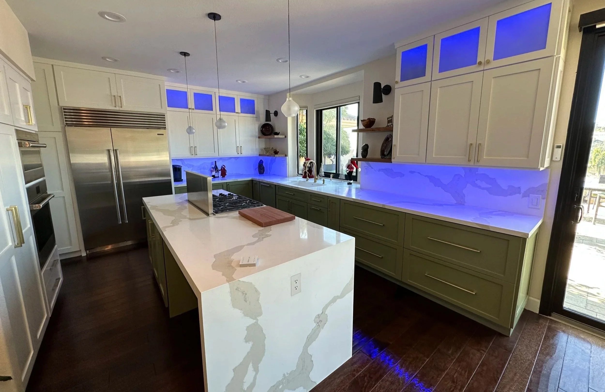 Modern kitchen with white and green cabinetry, a large white marble island with waterfall ends, stainless steel appliances, and blue LED lighting under the cabinets. There is a double oven, refrigerator, and large window at the sink.